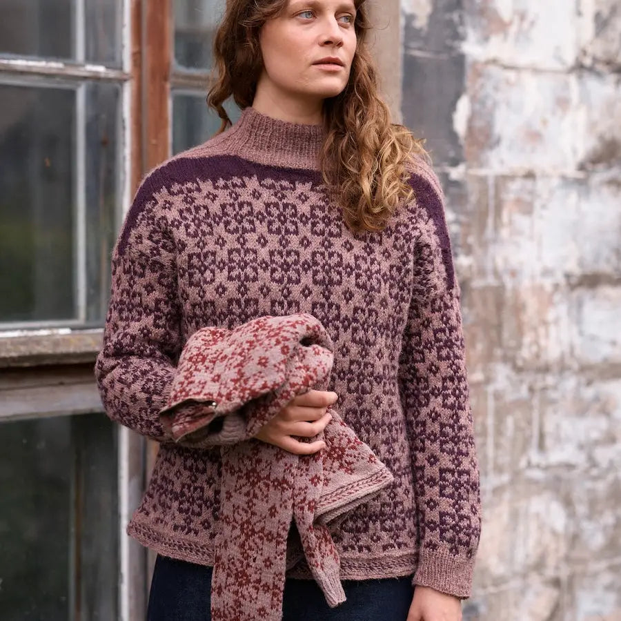Woman wearing a patterned sweater holding a scarf in front of a stone wall.