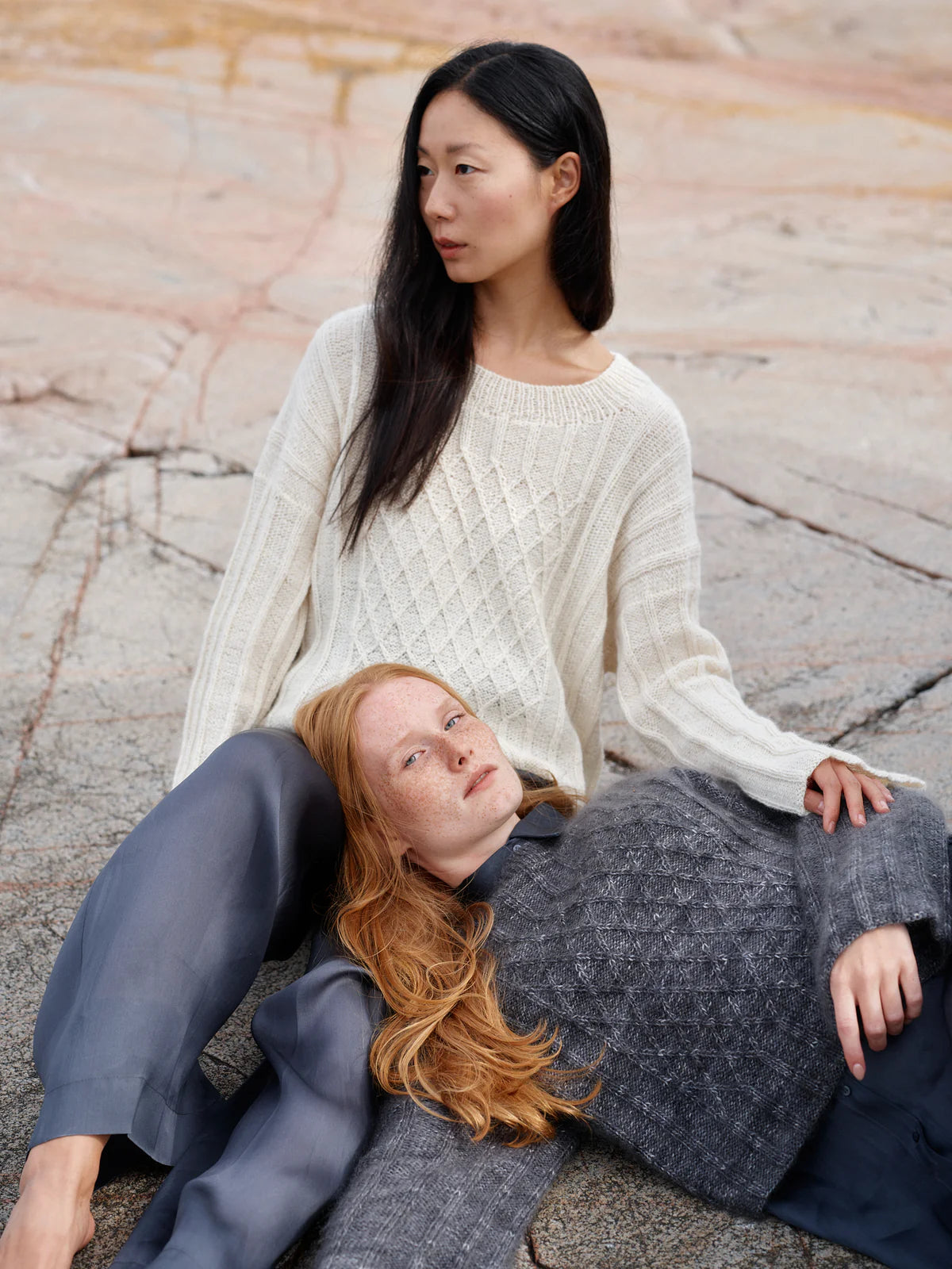 Two women sitting on a stone surface, one wearing a white sweater and the other in a gray outfit.