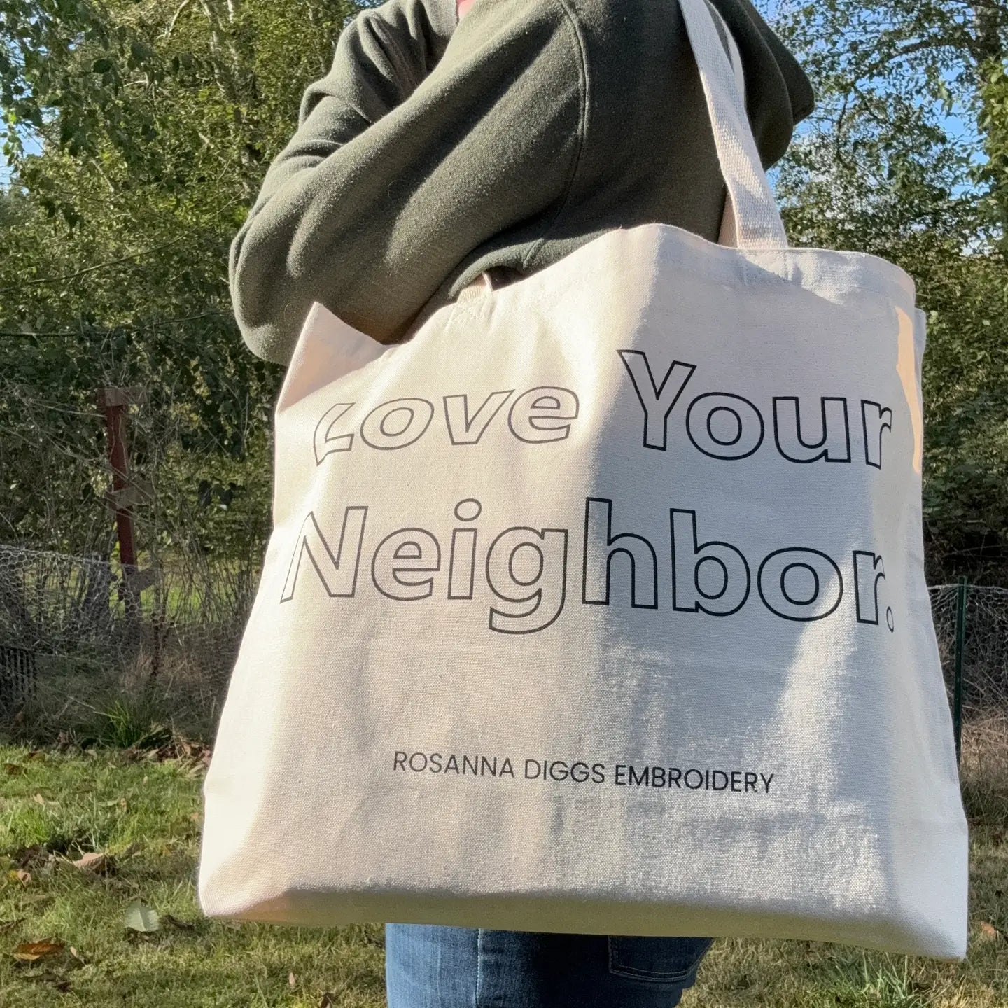 Person holding a tote bag with 'Love Your Neighbor' text and Rosanna Diggs Embroidery branding outdoors.