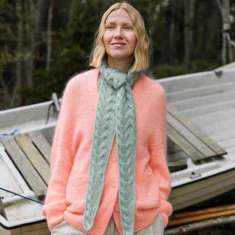 Woman wearing a pink sweater and green scarf standing near a boat in a forest setting