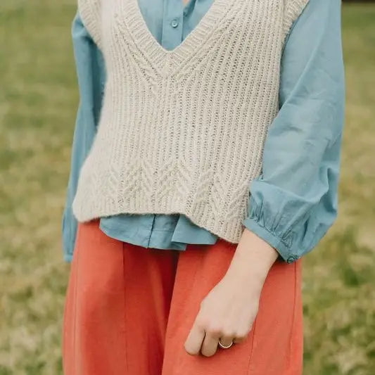 Person wearing a beige knitted vest over a blue shirt with red pants in a natural setting