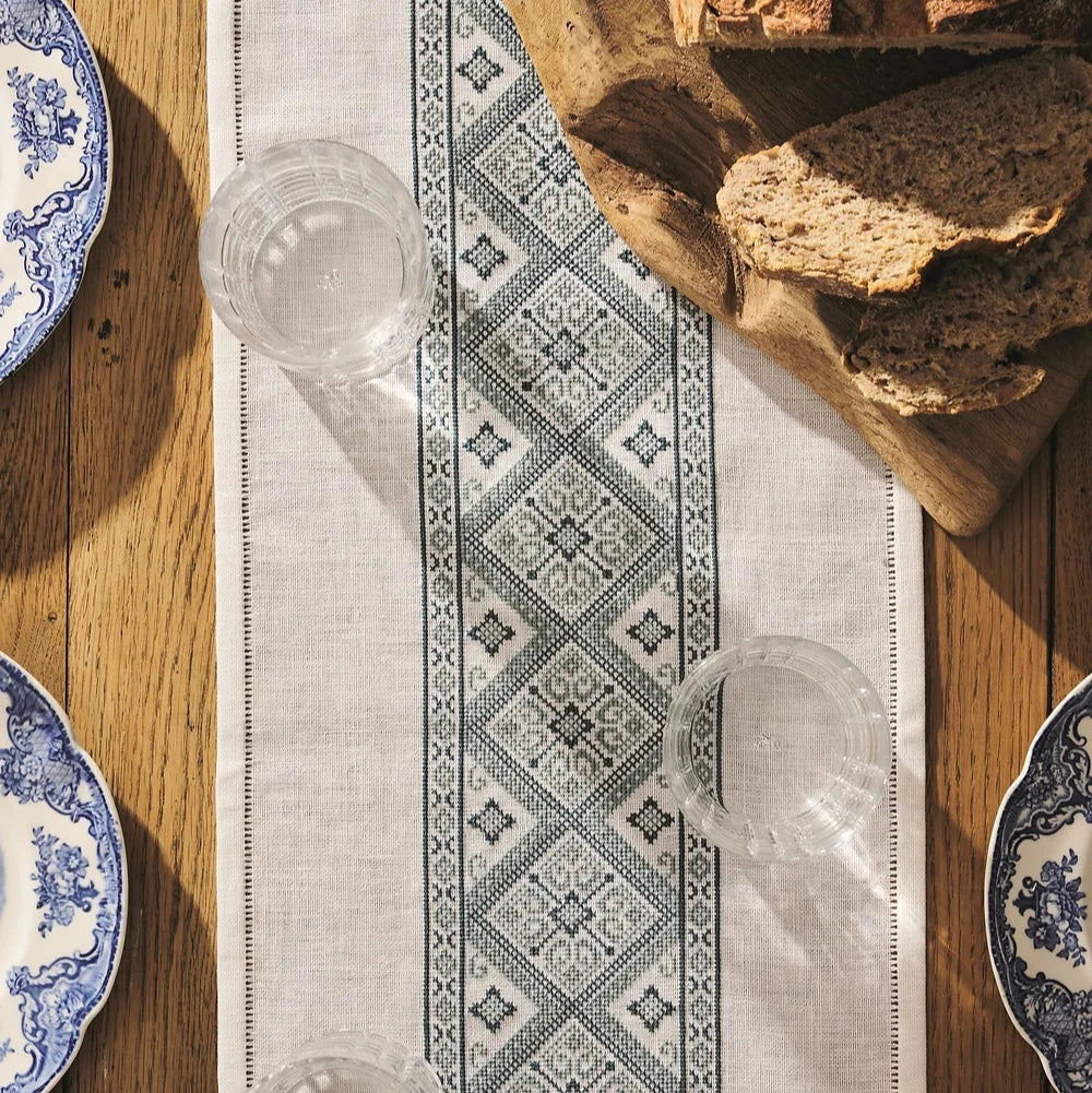 Cross stitch embroidery kit depicted with a linen fabric, floss, and tapestry needle, arranged on a table with a patterned plate and cutlery.