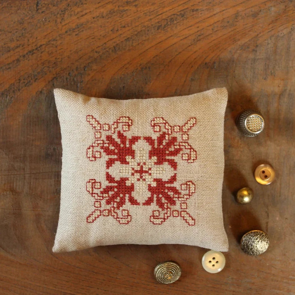 A cross-stitch embroidery pincushion kit, featuring a beige background with a red and dark beige star design, displayed on a wooden surface with buttons and thread around it.