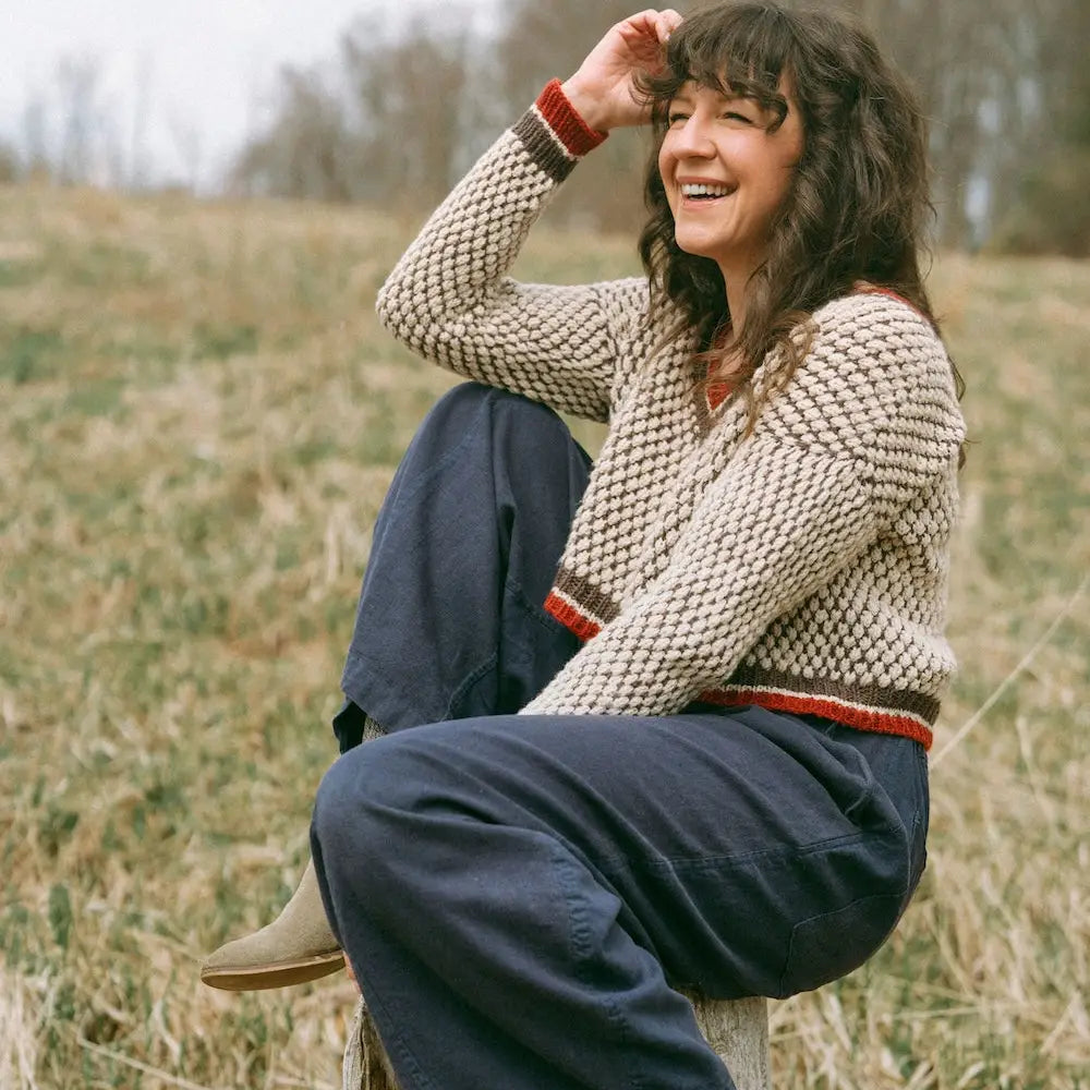 Woman sitting outdoors wearing a chunky knit sweater and blue pants.