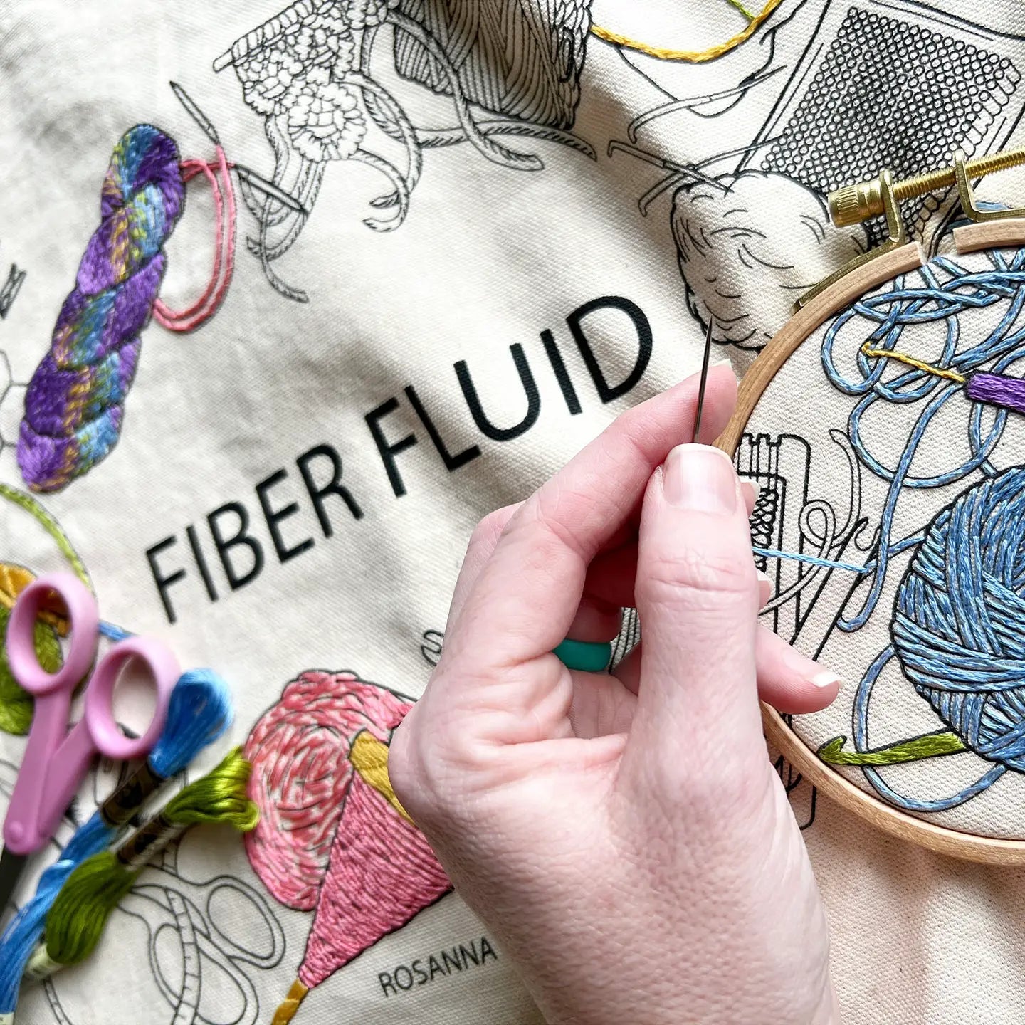 Hand embroidery with thread and tools on a surface with 'Fiber Fluid' branding.