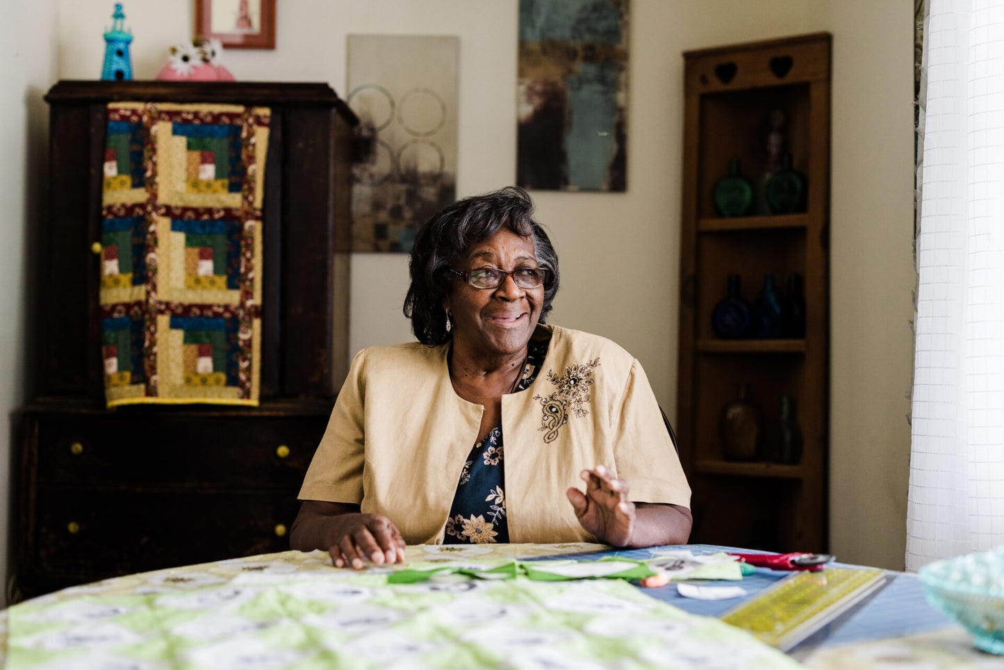 Woman sitting at a table with quilting materials, smiling in a home setting.
