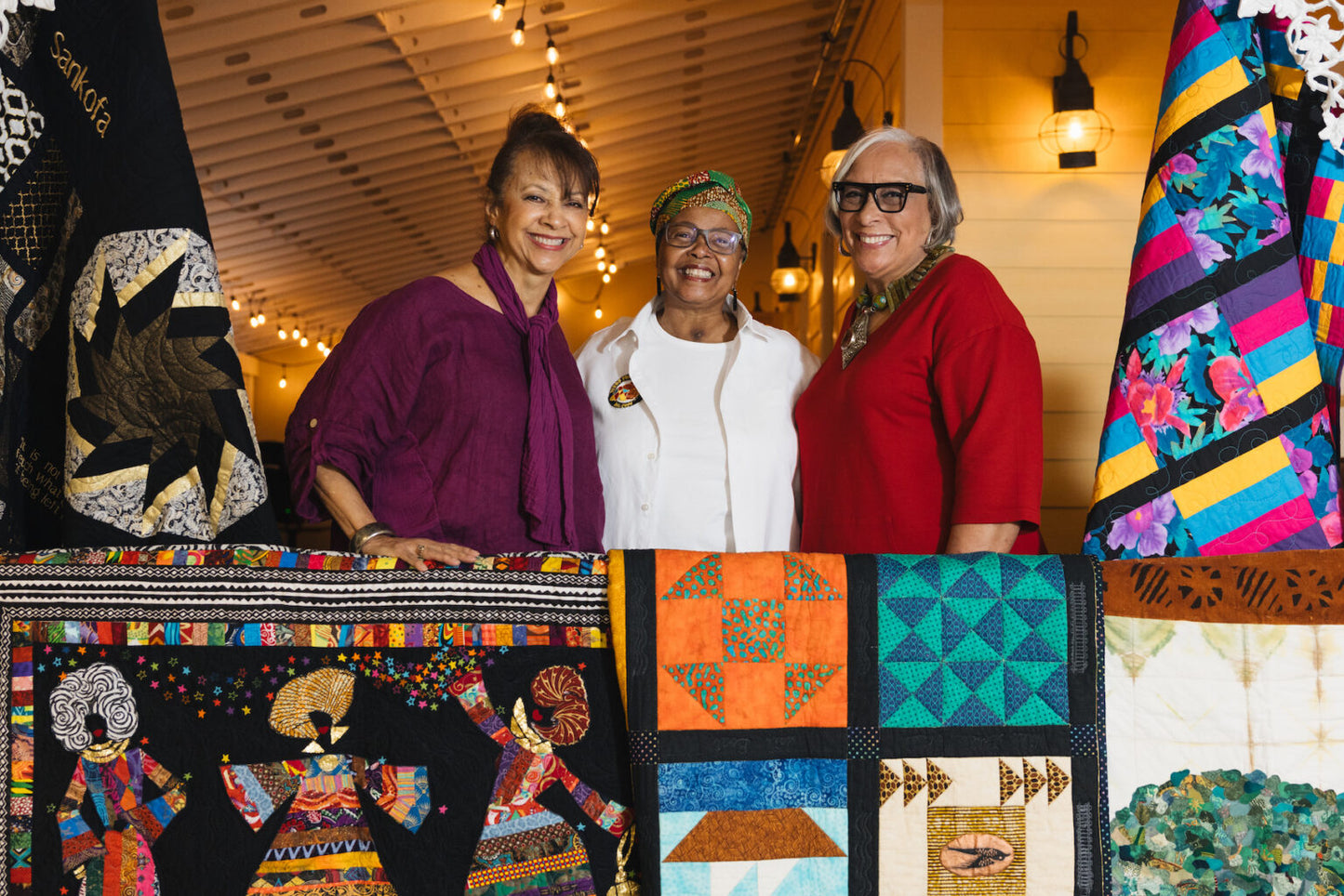 Three women standing behind a display of colorful quilts in a store setting.