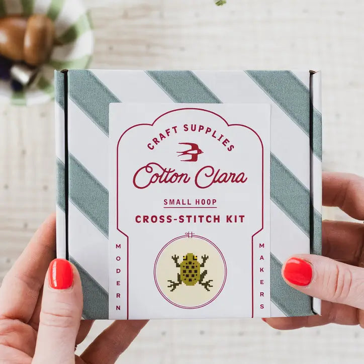 Cotton Clara cross-stitch kit held by a person with red nail polish on a light background.