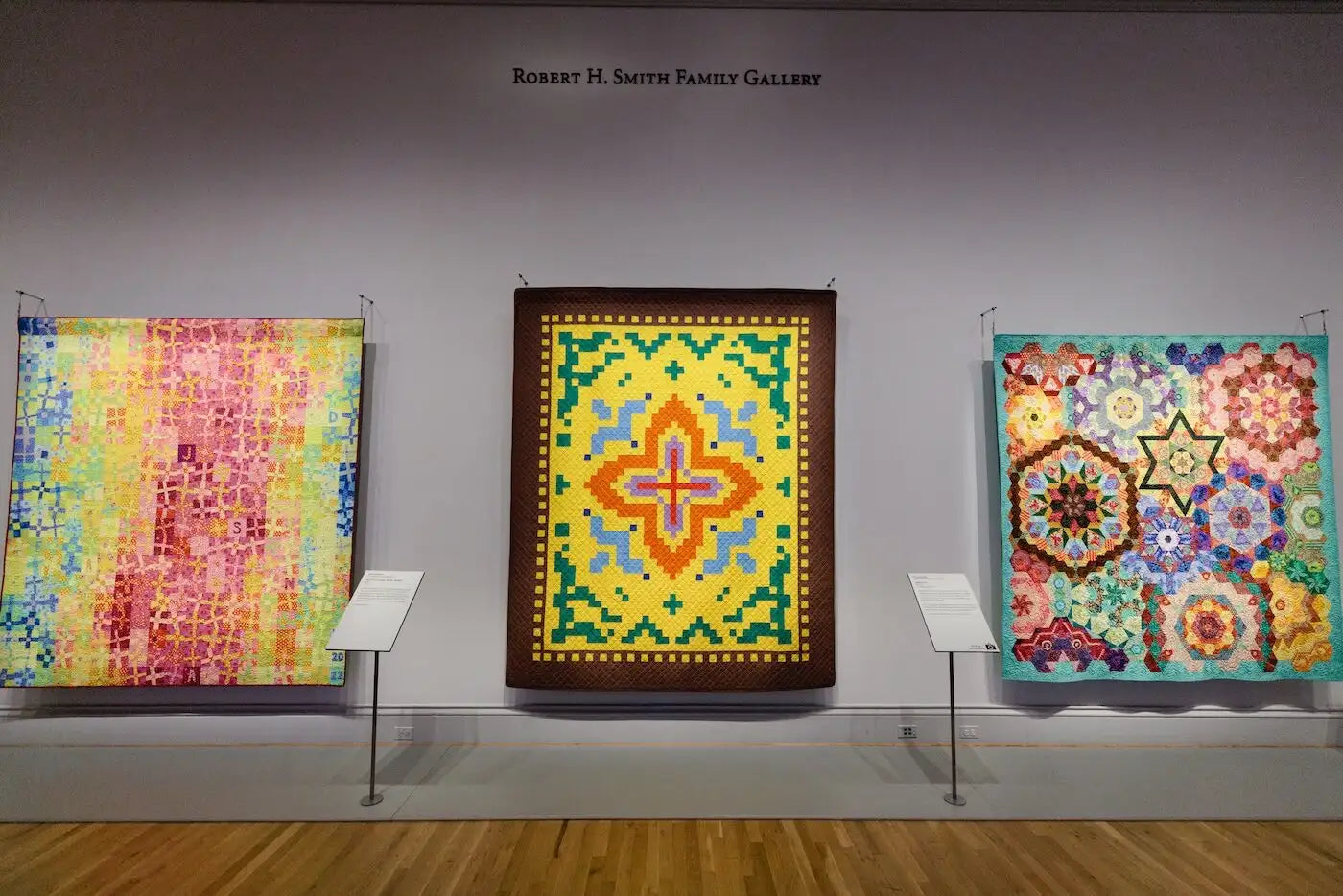 Three colorful quilted wall hangings displayed in a gallery setting. A central framed quilt with a bright yellow and orange geometric star design hangs in the middle, flanked by two more patterned quilts in pink, blue, and multicolor tones.