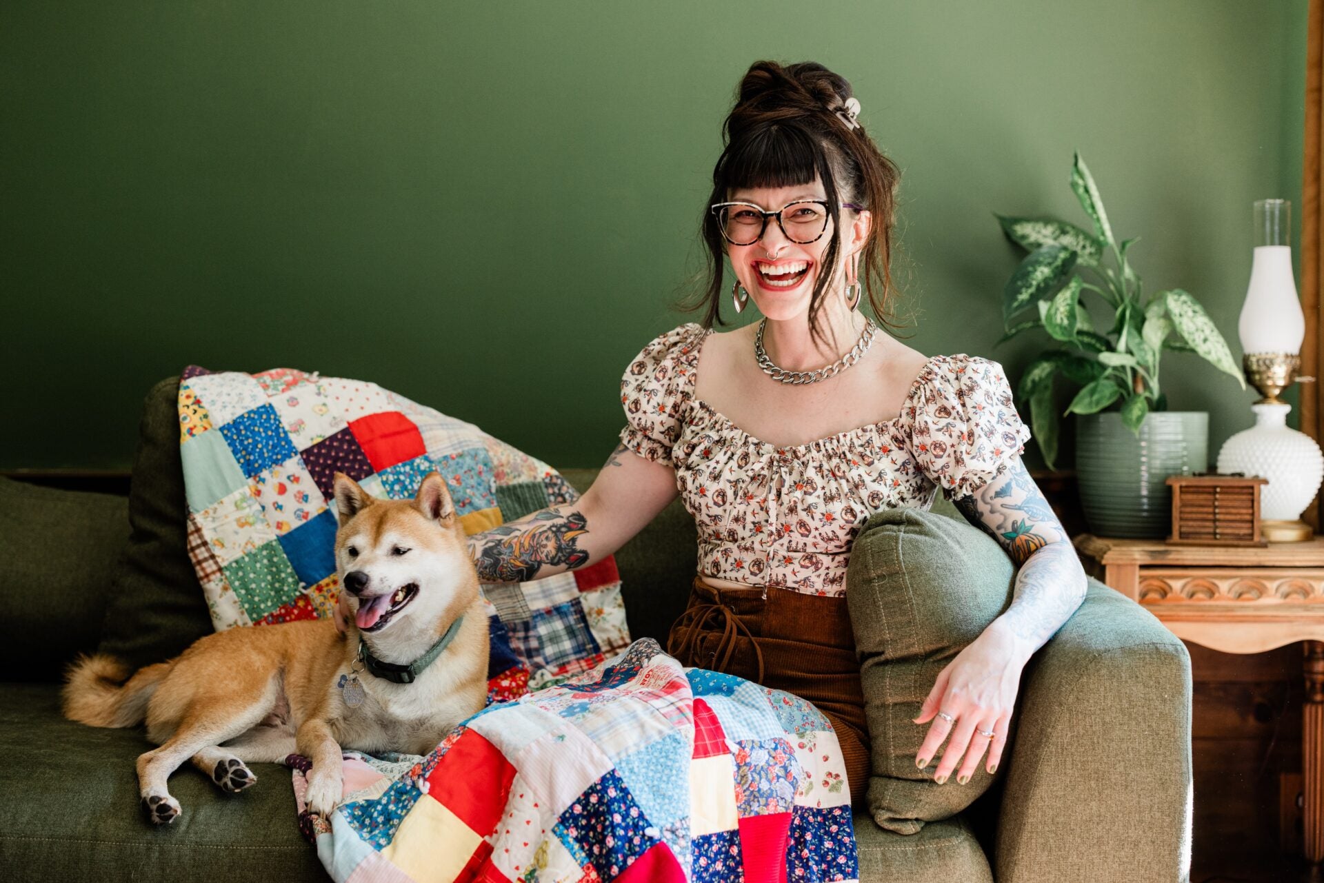Woman sitting on a couch with a quilt and a dog, surrounded by decorative pillows and plants.