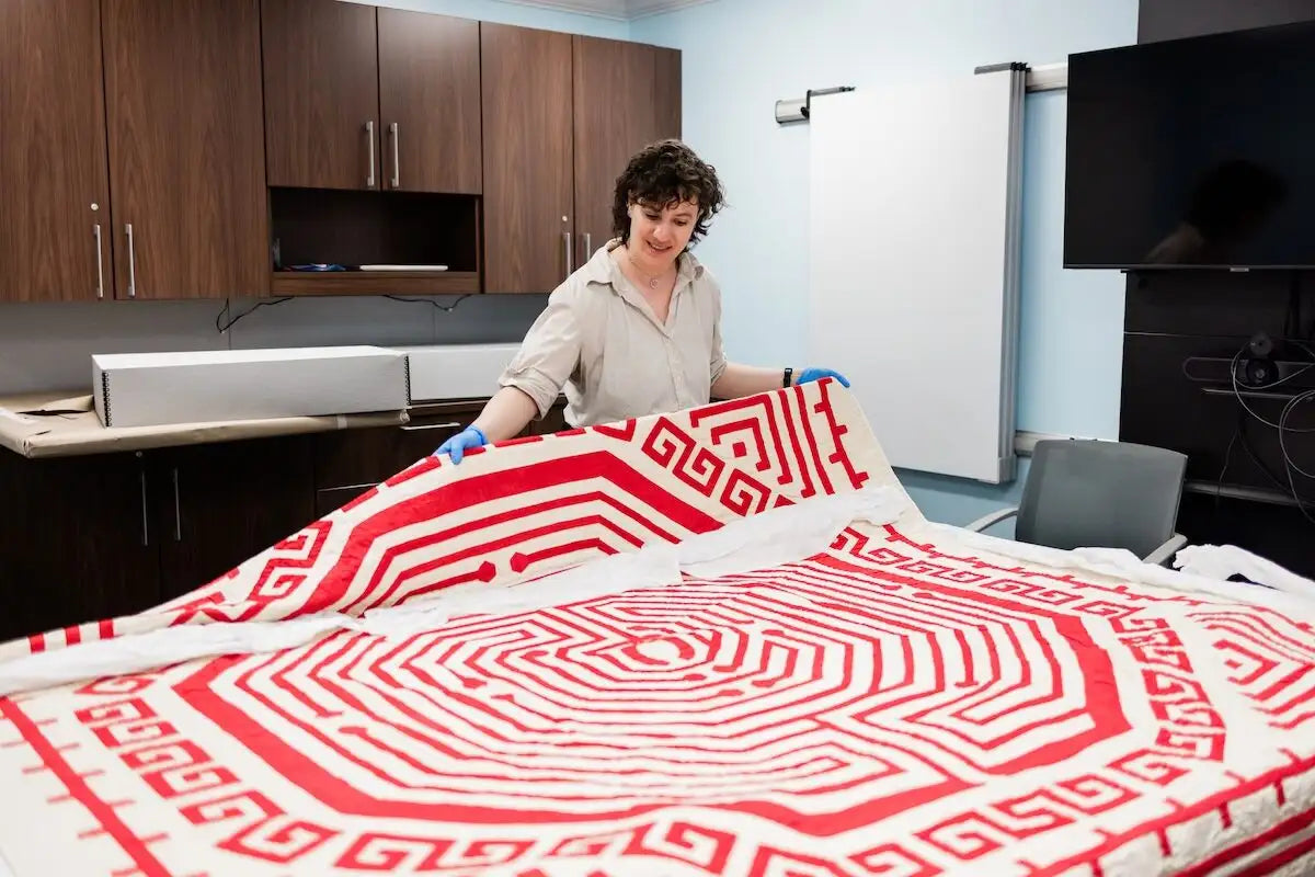 A person wearing blue gloves holds up a large white textile with a bold red geometric maze-like pattern 