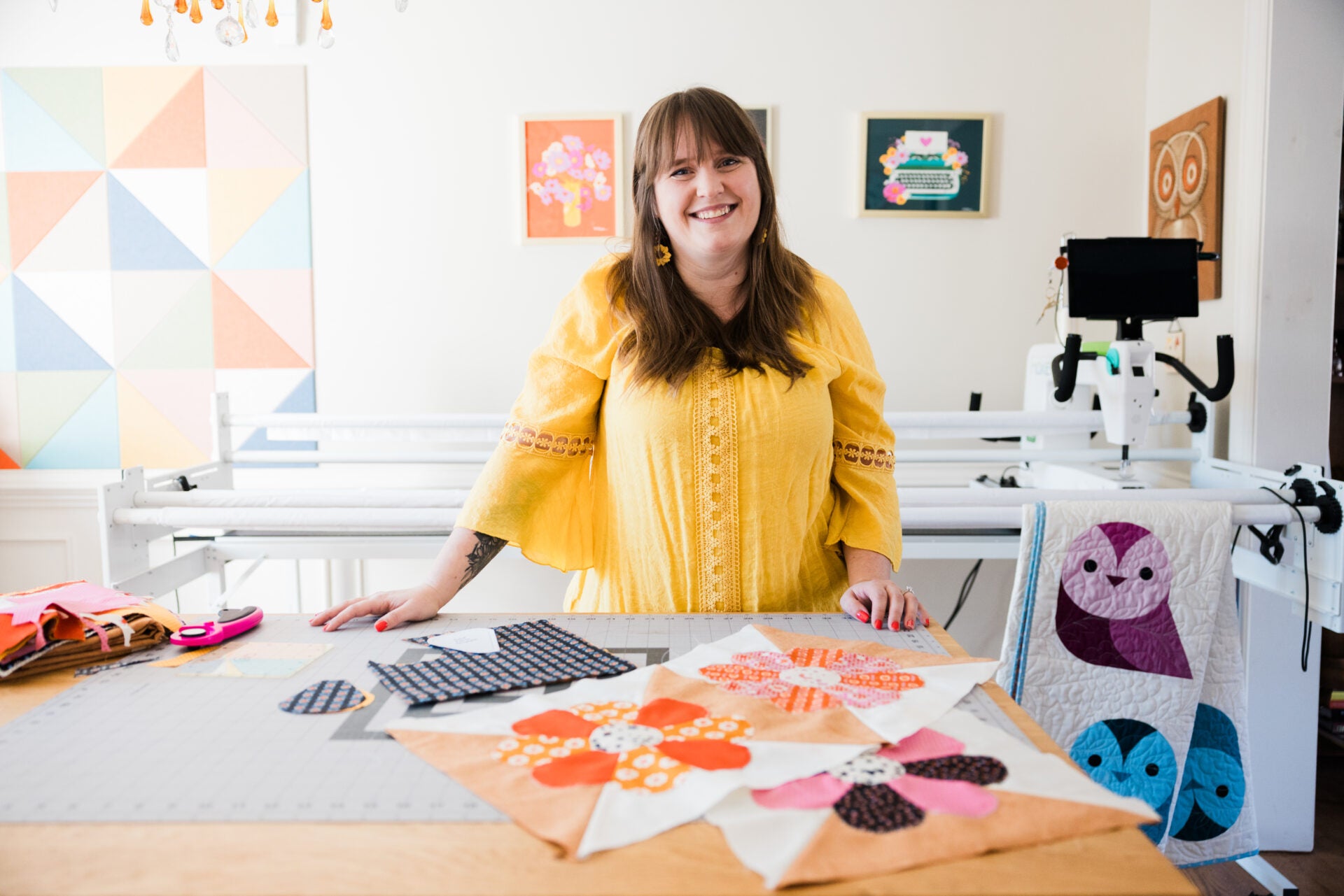Woman in a yellow shirt standing in front of a table with quilt block samples and sewing equipment.