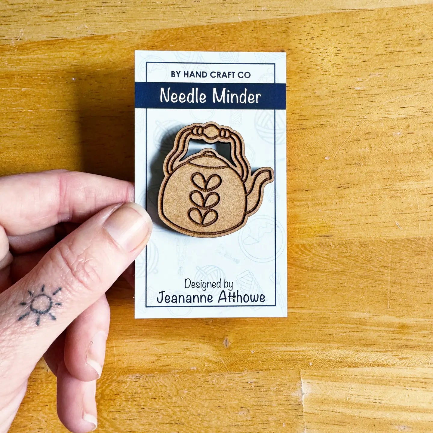 Wooden needle minder on a card held by a hand with a tattoo on a wooden surface