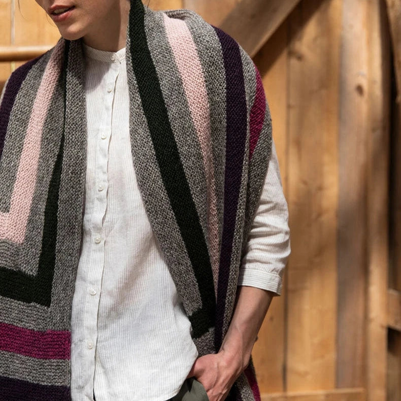 Woman wearing a patterned cardigan over a white shirt and green pants, standing against a wooden wall.