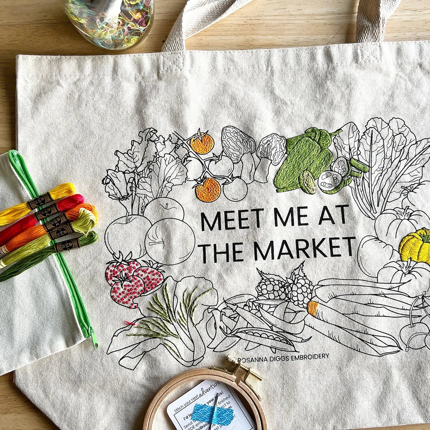 Embroidery hoop with embroidered vegetables and text 'Meet me at the market' on a white fabric background.