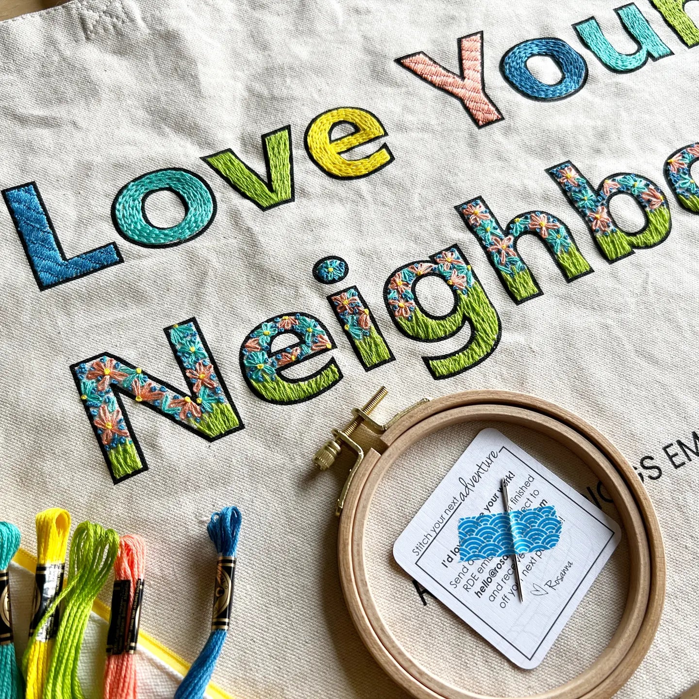 Embroidery hoop with thread and a card on a fabric background with colorful text.