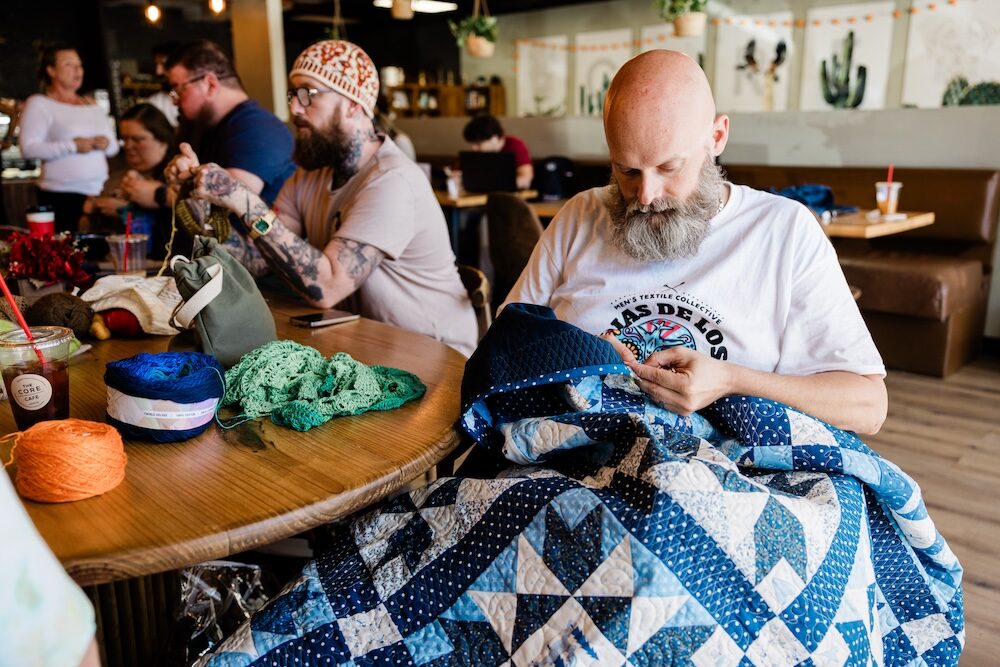 Man working on a quilt in a casual setting with people and tables in the background
