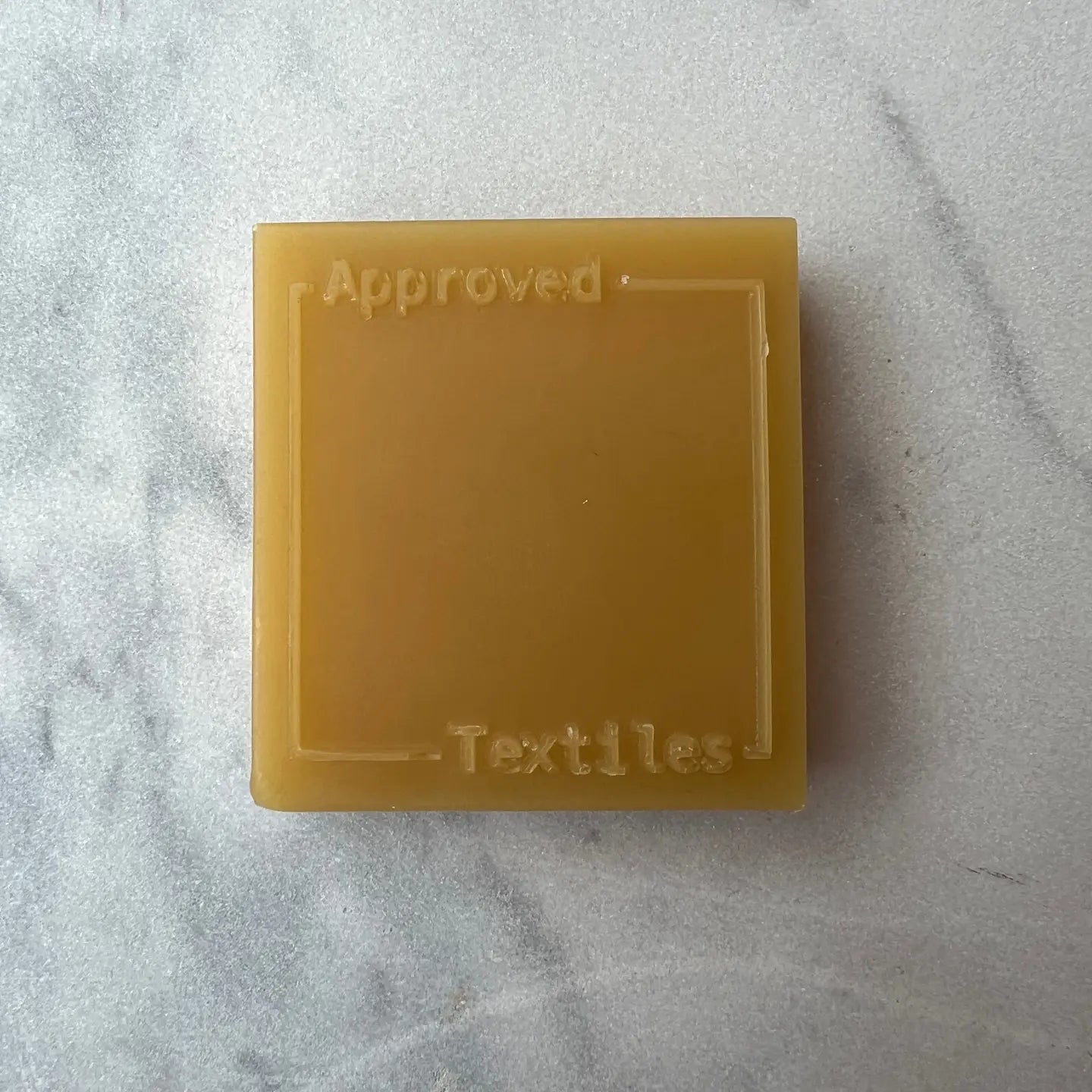 Yellow square beeswax block with 'Approved Textiles' text on a gray marble surface