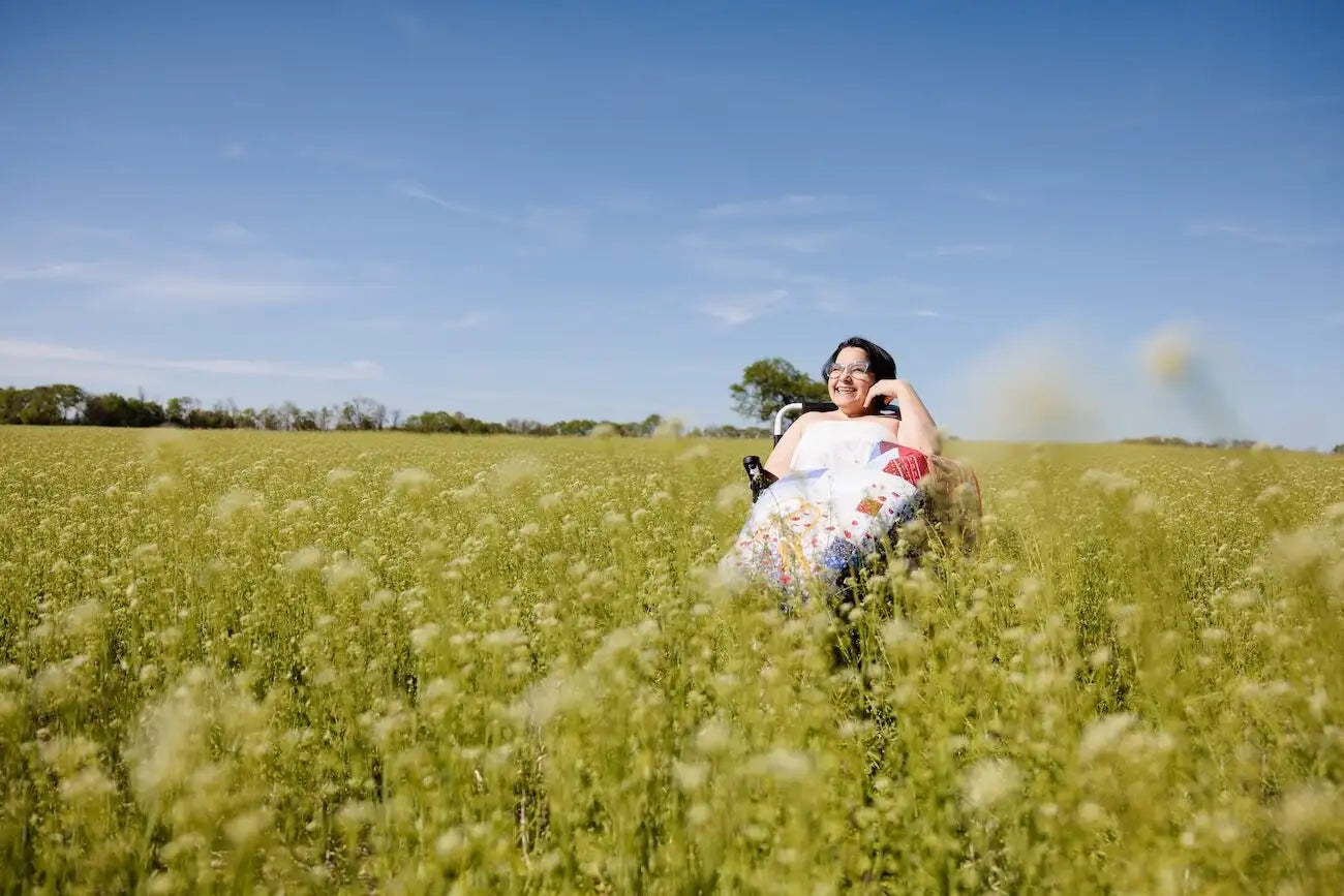 A woman sitting in a wheelchair in a field of green plants under a blue sky, wearing a white sleeveless top and a floral skirt.