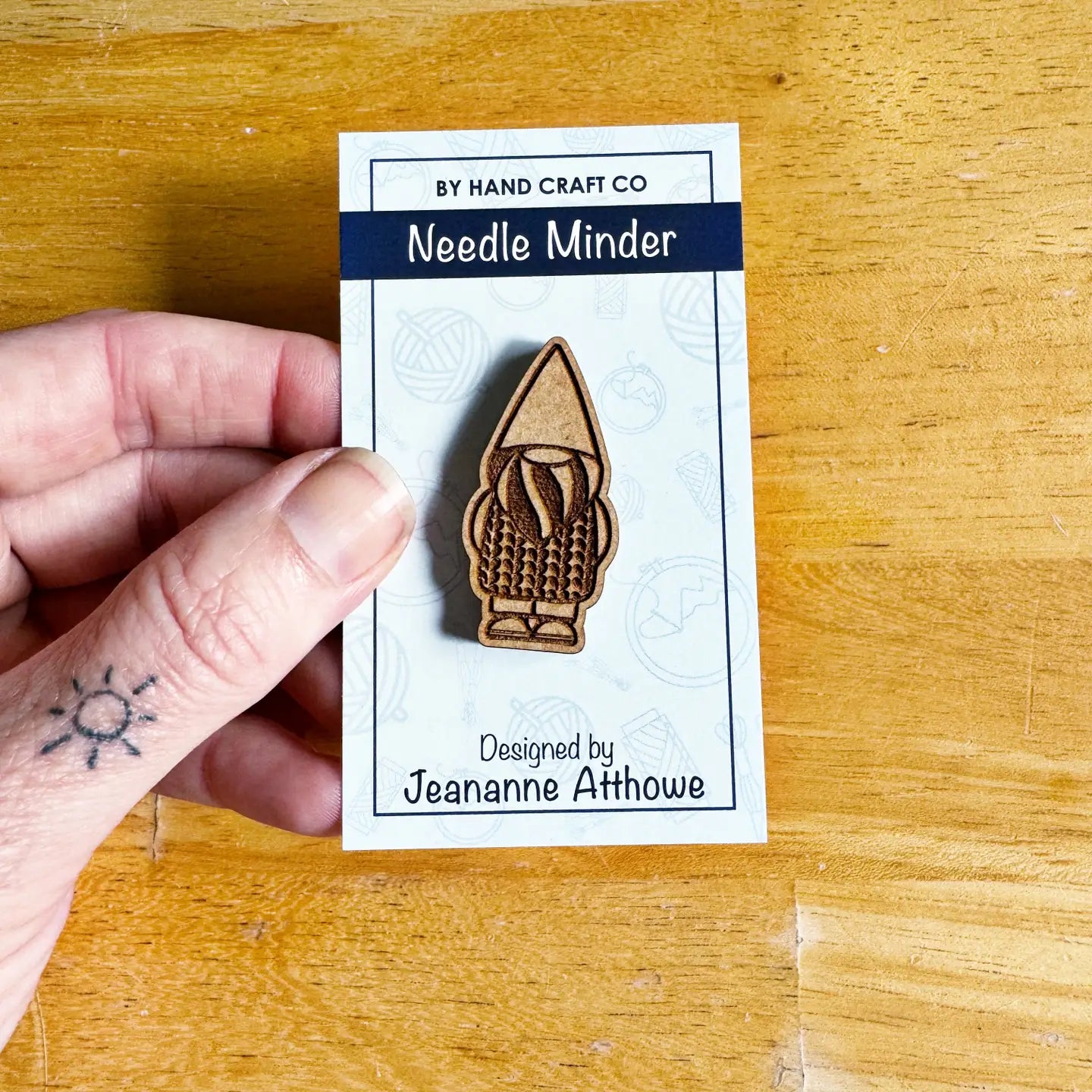 Hand holding a needle minder on a By Hand Craft Co branded card with 'Designed by Jeananne Aithowe' text.