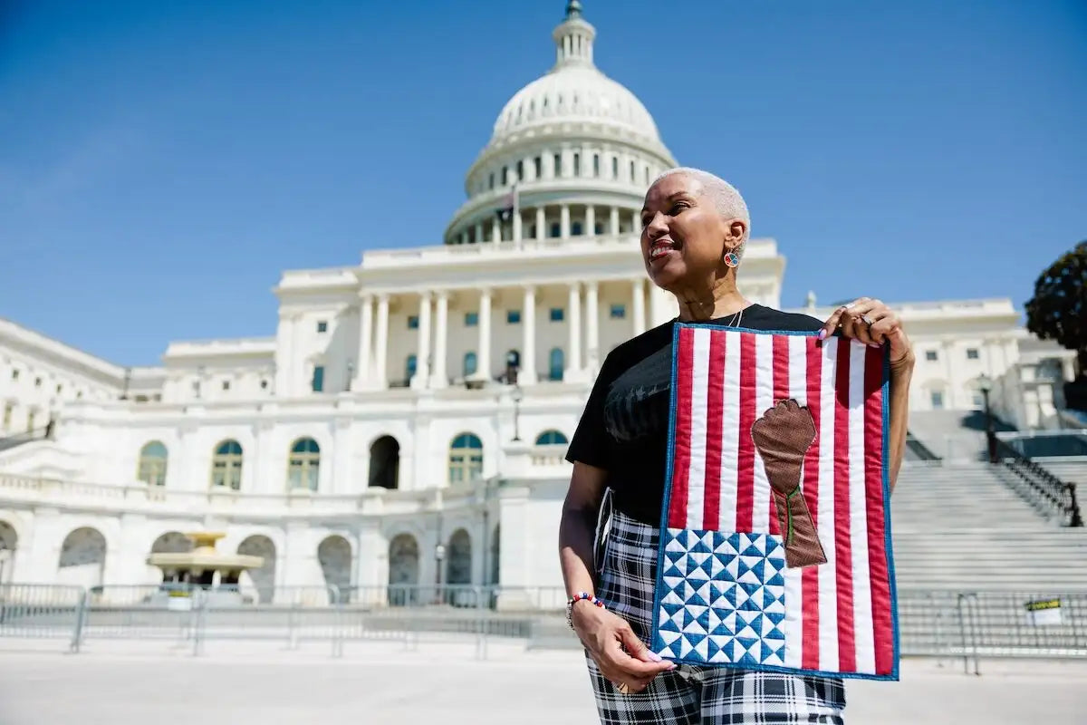 A person stands outdoors in front of the U.S. capitol with a small flag quilt