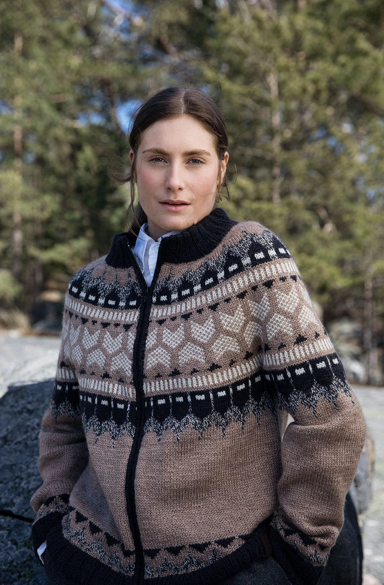 A woman wearing a brown and black patterned knit zip-up sweater outdoors in front of trees.