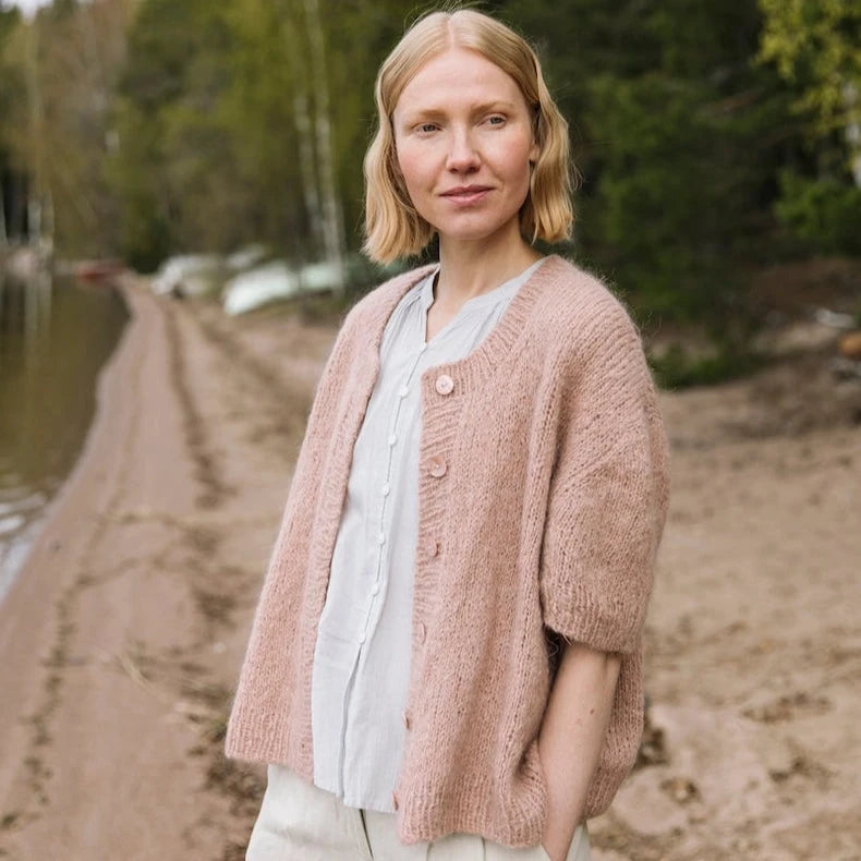 Woman wearing a pink cardigan standing on a dirt path with trees in the background