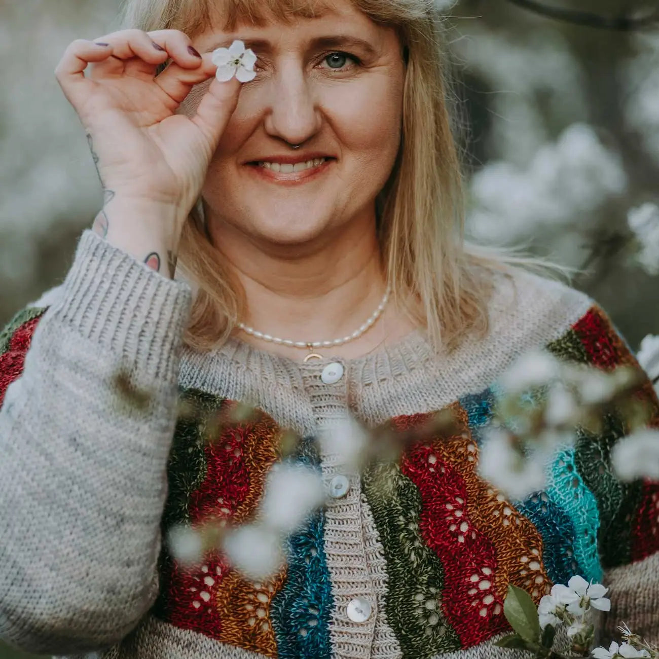 Woman holding a white flower near her eye with a colorful sweater in a natural setting
