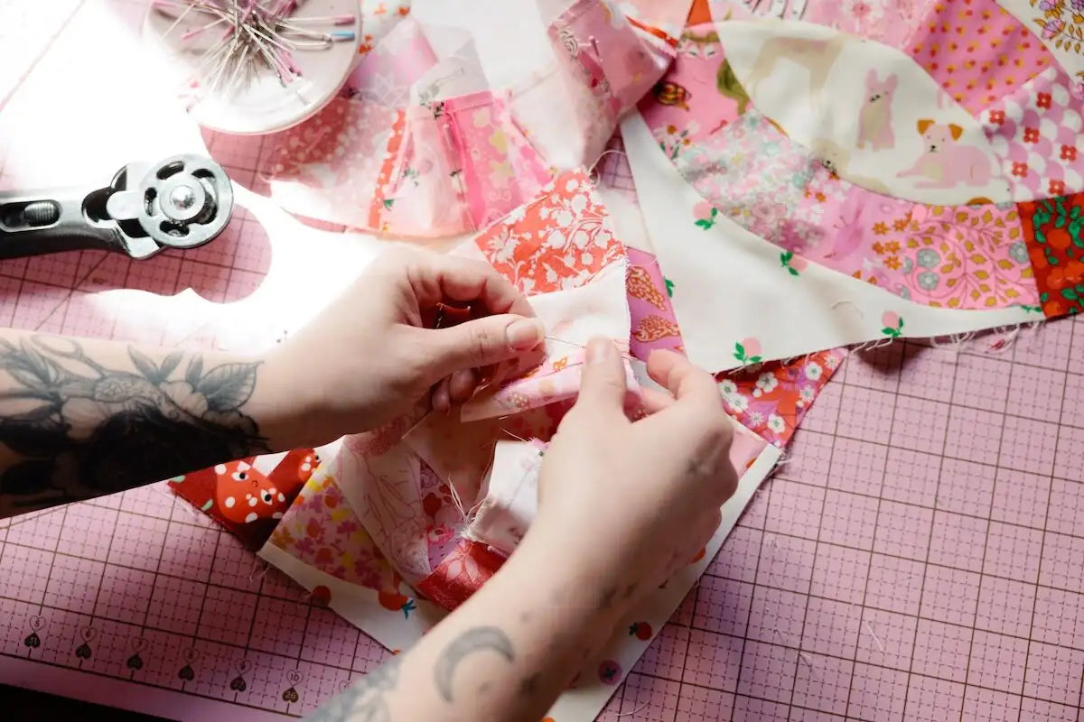 A close-up of a person's tattooed hands folding a piece of pink and white patterned fabric on a pink cutting mat, with rotary cutters and other fabric pieces visible in the background.