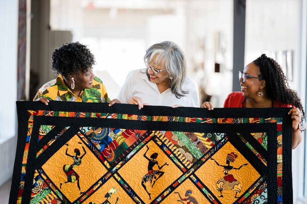 Three women holding a colorful quilt with African-inspired designs.