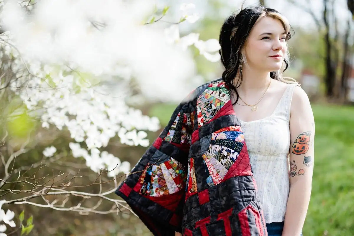 A woman standing outdoors in a grassy park next to a flowering tree, wearing a colorful patchwork quilt draped over her shoulders and a white sleeveless top.