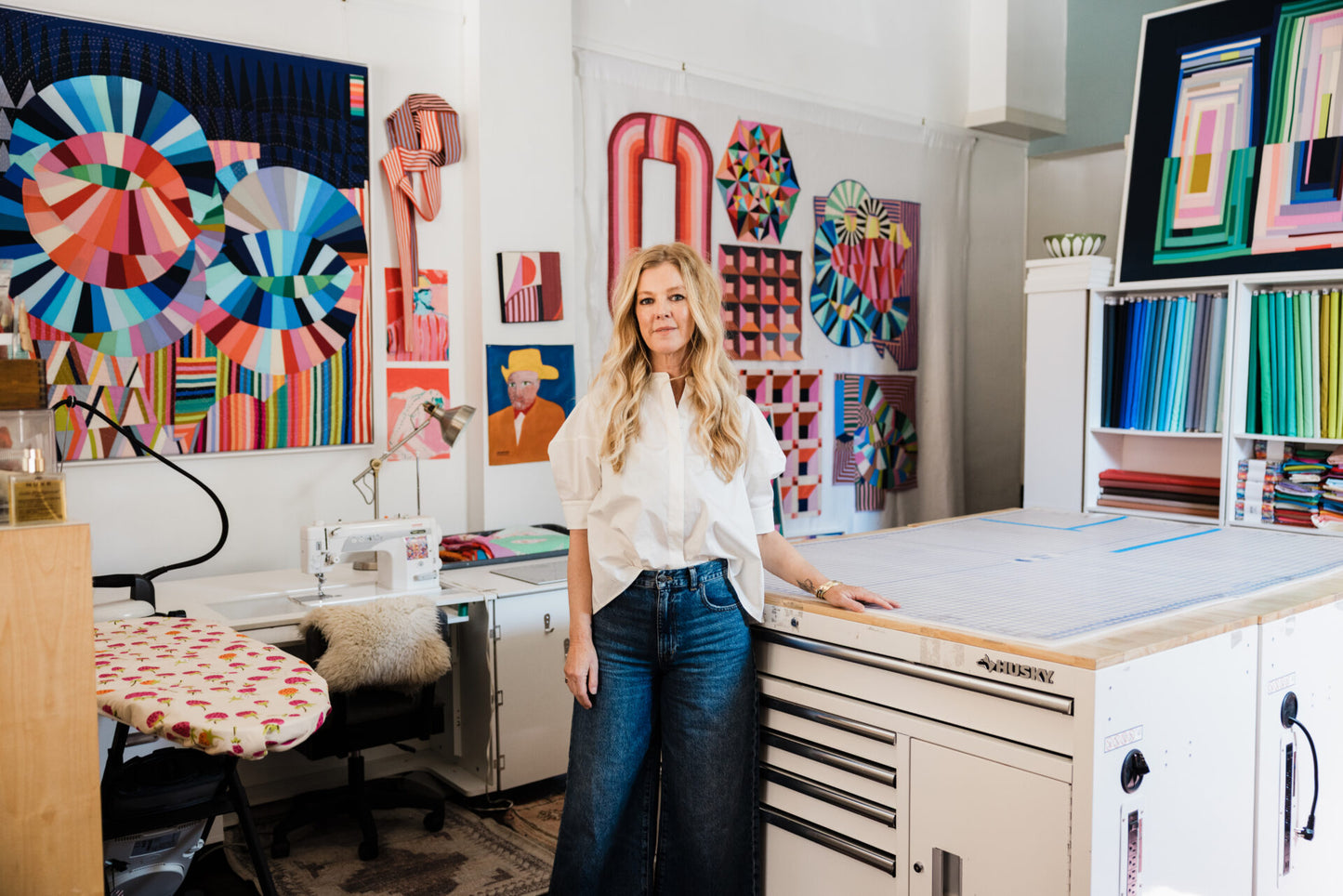 Woman standing in a creative workspace with colorful quilts and sewing equipment.