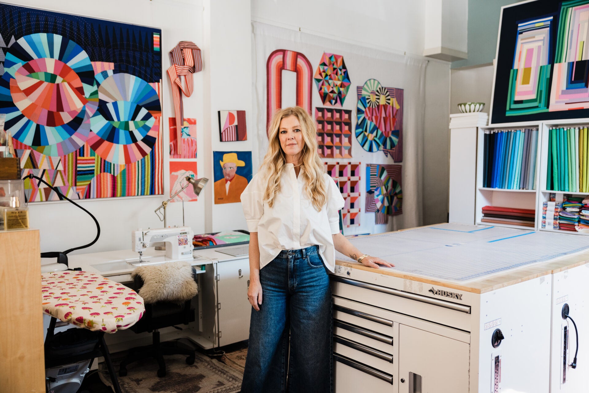 Woman standing in a creative workspace with colorful quilts and sewing equipment.