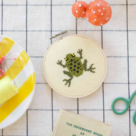 Embroidery hoop with a frog design on a checkered tablecloth with sewing tools.