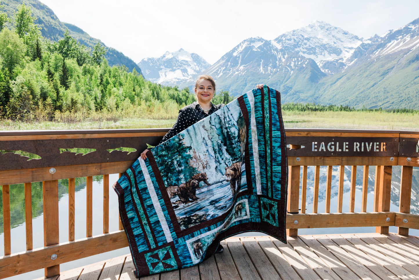 Person holding a quilt with nature design in front of mountains and water