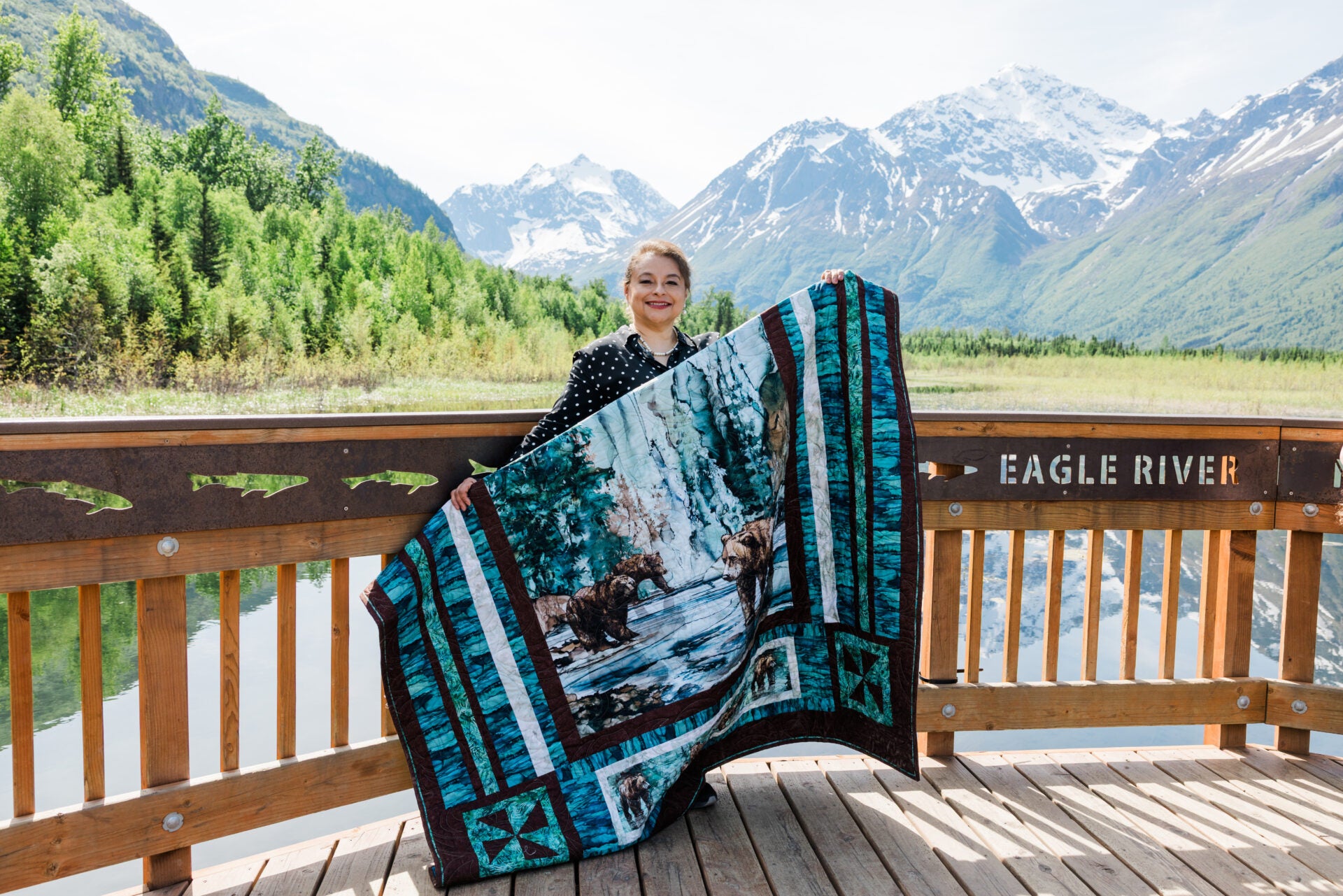 Person holding a quilt with nature design in front of mountains and water
