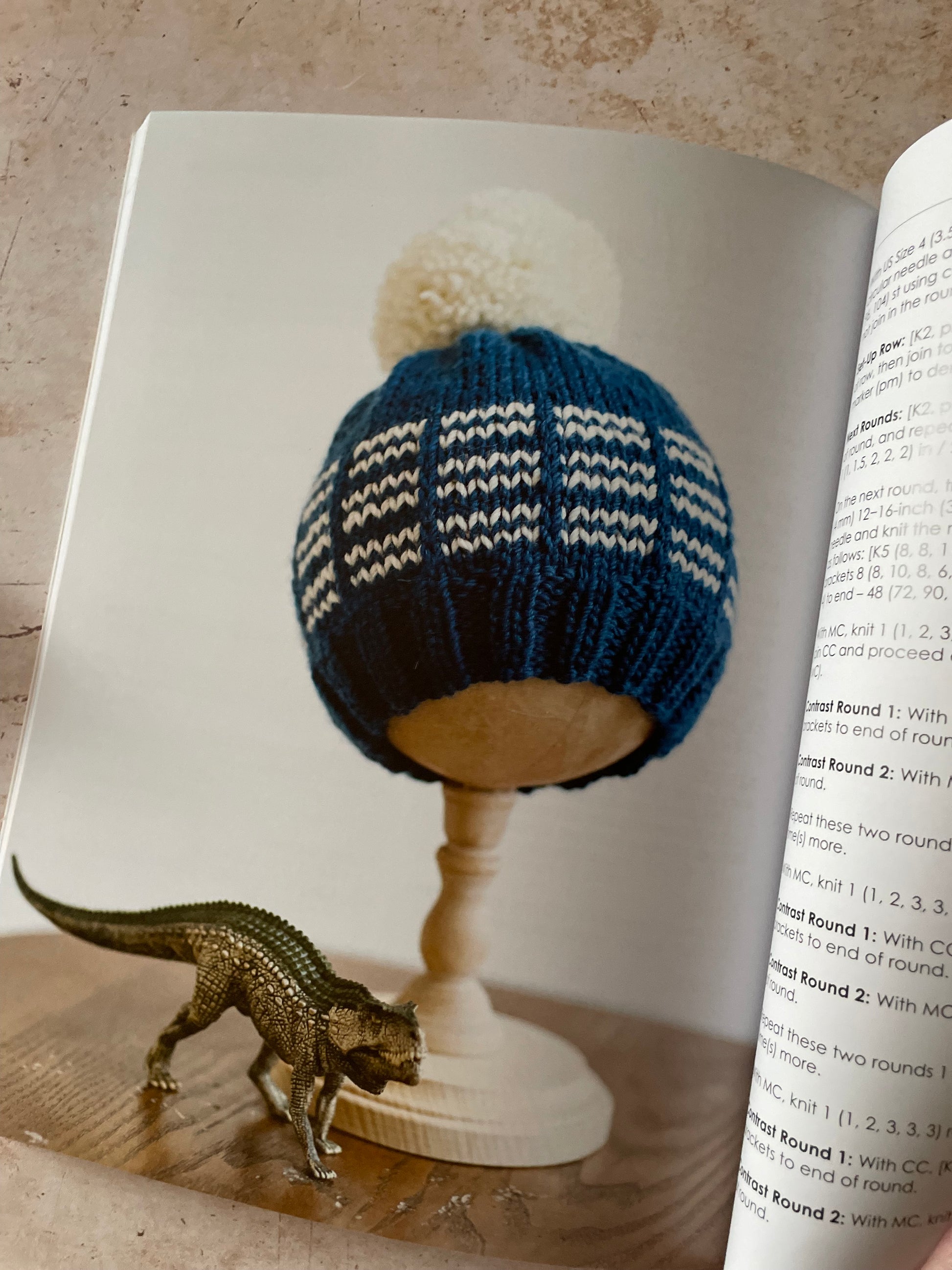 Blue knitted hat with white stripes on a wooden hat stand, with a dinosaur in the foreground