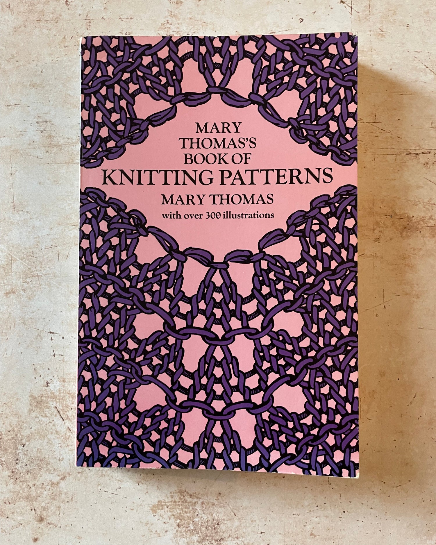 Book cover of 'Mary Thomas's Book of Knitting Patterns' with a pink and purple design on a textured beige background.