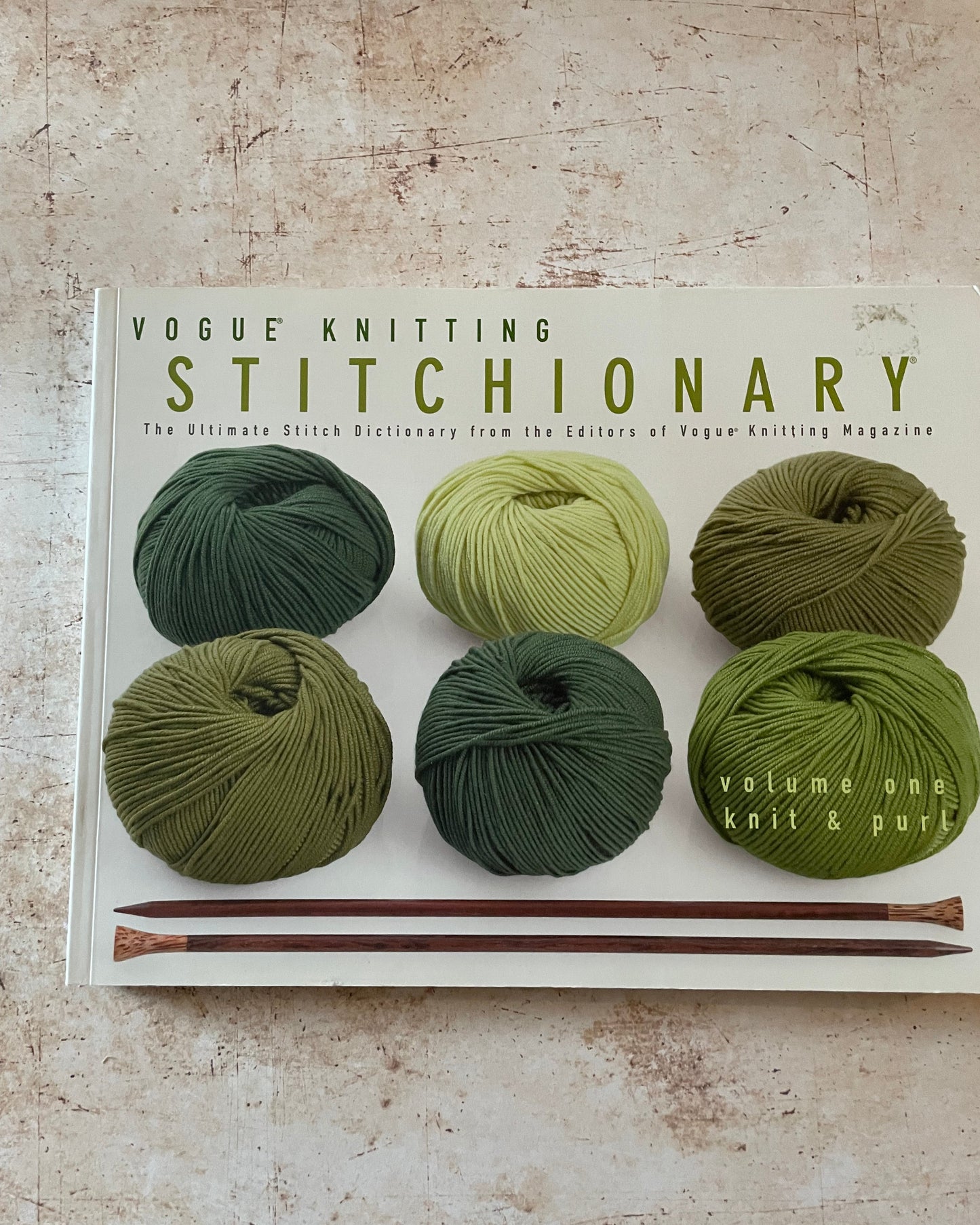 Book titled 'Vogue Knitting Stitchionary' with green yarn and knitting needles on a textured surface