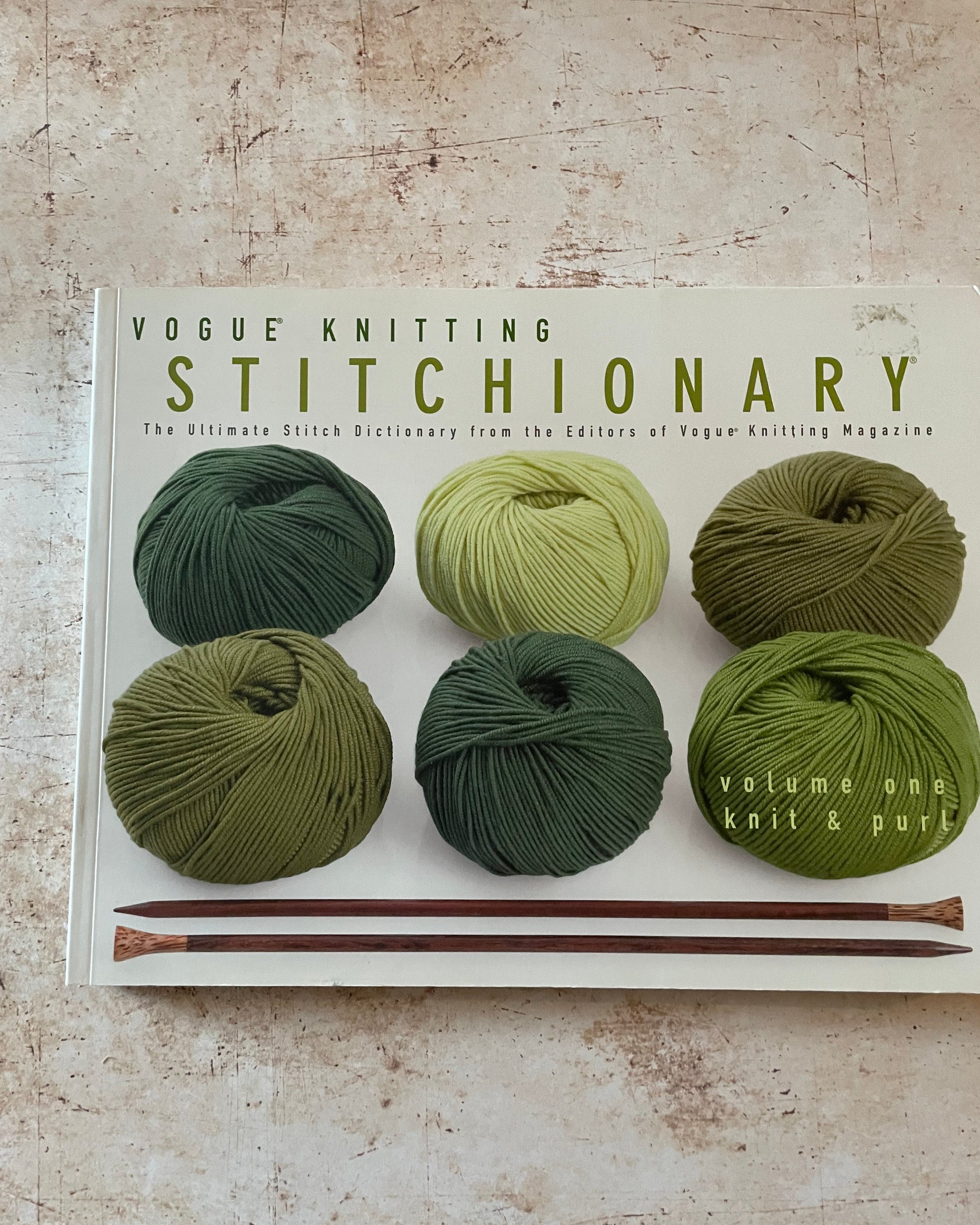 Book titled 'Vogue Knitting Stitchionary' with green yarn and knitting needles on a textured surface