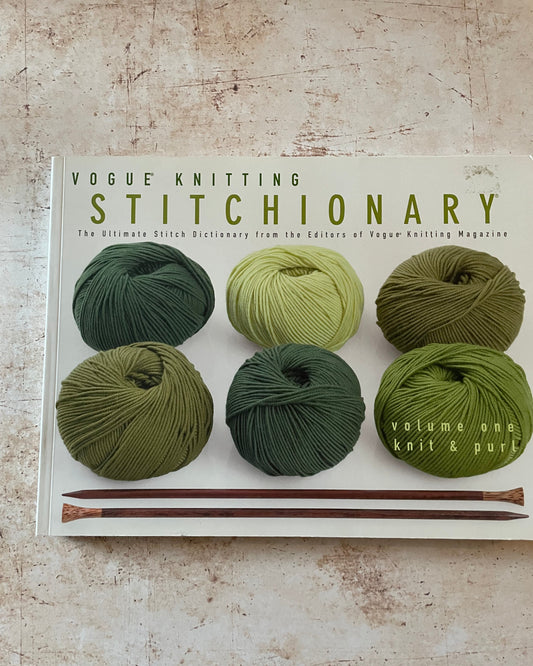 Book titled 'Vogue Knitting Stitchionary' with green yarn and knitting needles on a textured surface
