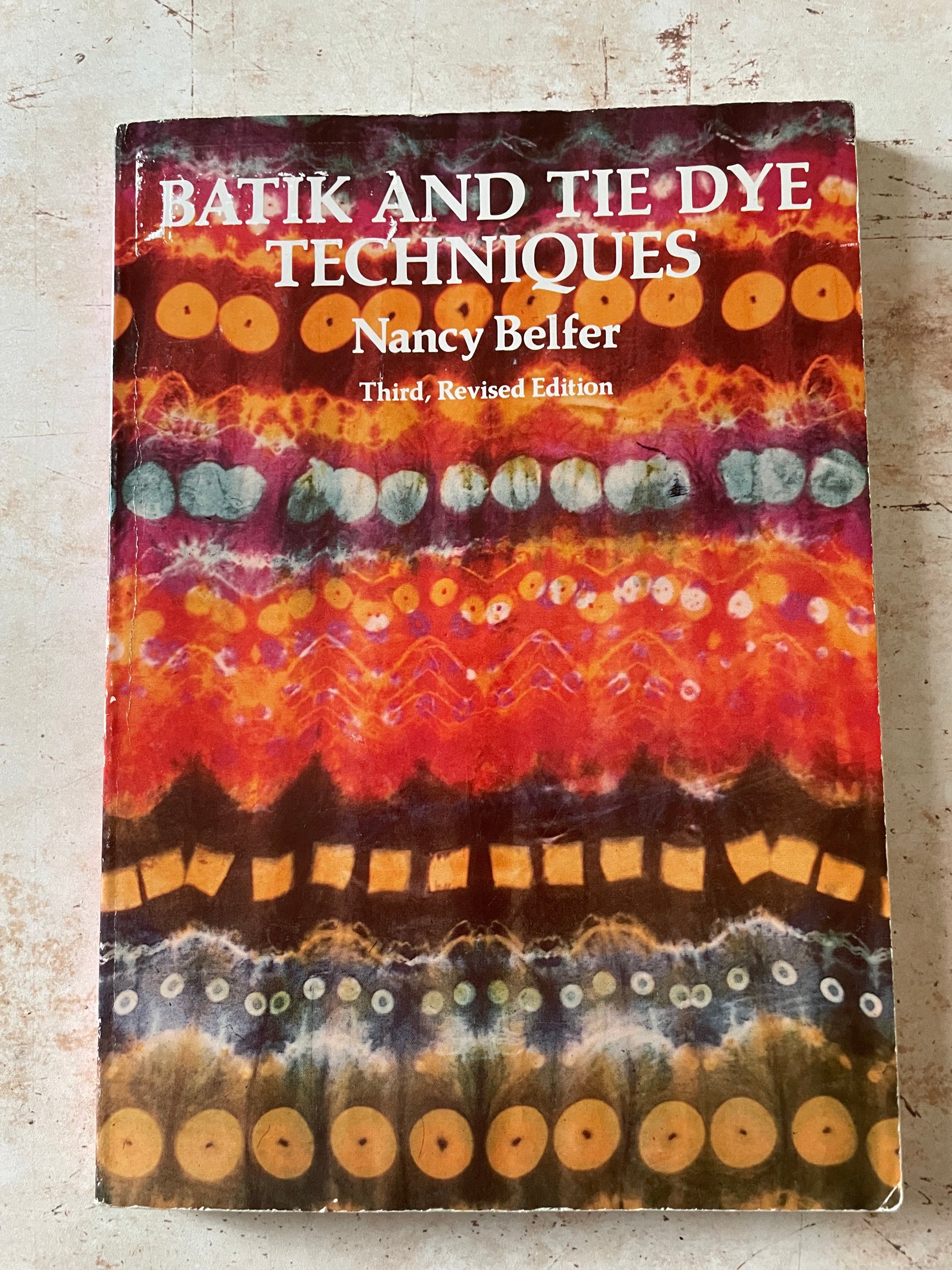 A book titled 'Batik and Tie Dye Techniques' by Nancy Belker, showing a colorful cover with batik patterns in red, orange, yellow, and brown on a light textured surface.