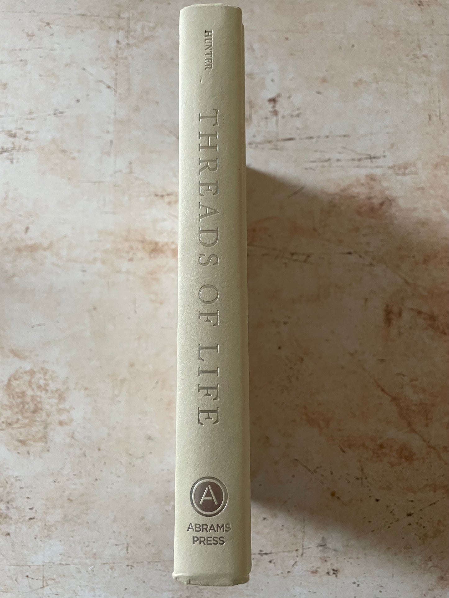 The spine of the book 'Threads of Life' by Hunter, published by Abrams Press, standing vertically on a textured beige stone surface.