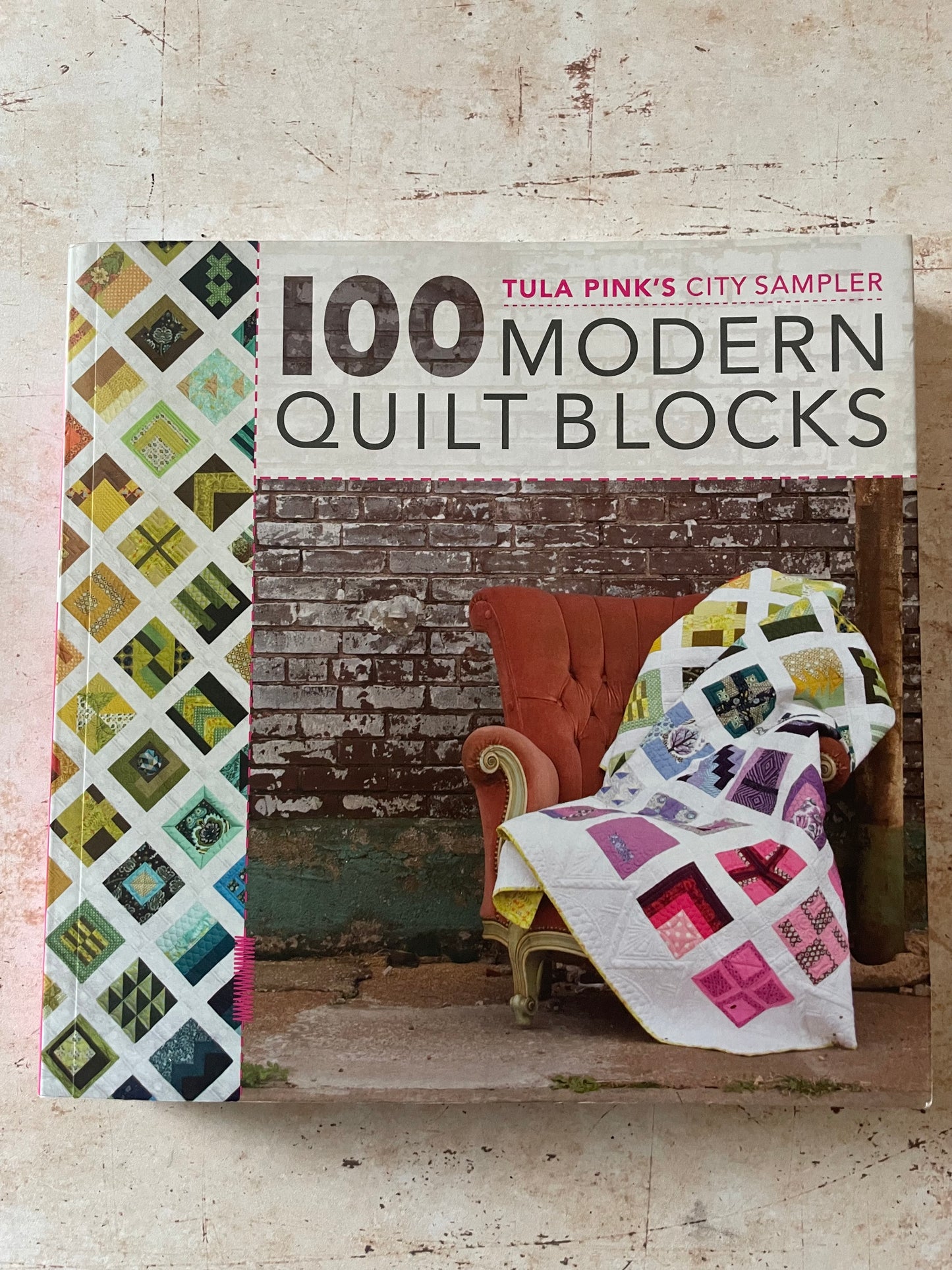 The front cover of the book '100 Modern Quilt Blocks' by Tula Pink, City Sampler, displayed on a textured beige surface. The cover features a colorful quilt pattern on the left, a brick wall background, and an orange armchair draped with a patchwork quilt on the right.