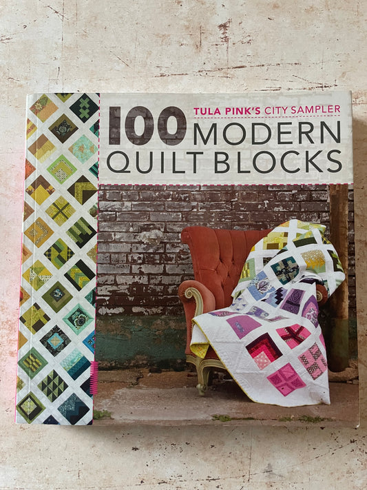 The front cover of the book '100 Modern Quilt Blocks' by Tula Pink, City Sampler, displayed on a textured beige surface. The cover features a colorful quilt pattern on the left, a brick wall background, and an orange armchair draped with a patchwork quilt on the right.