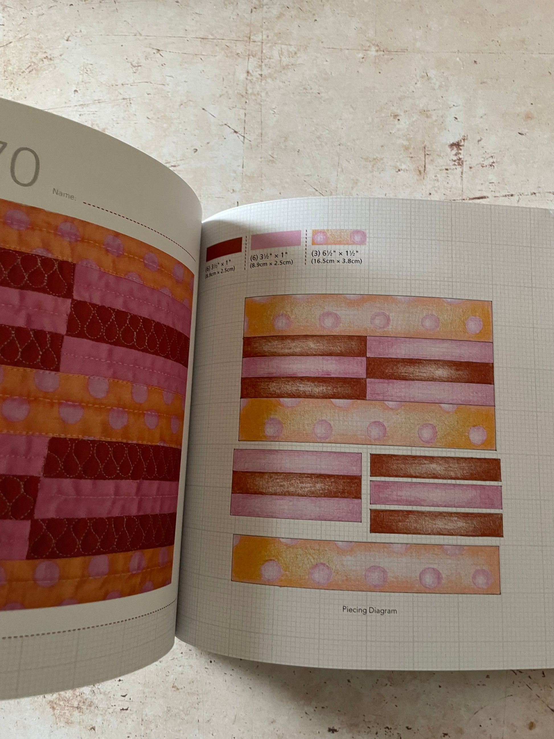 An open quilt pattern book showing sewing and quilting diagrams with colorful fabric swatches in shades of pink, orange, and red on a beige stone surface.