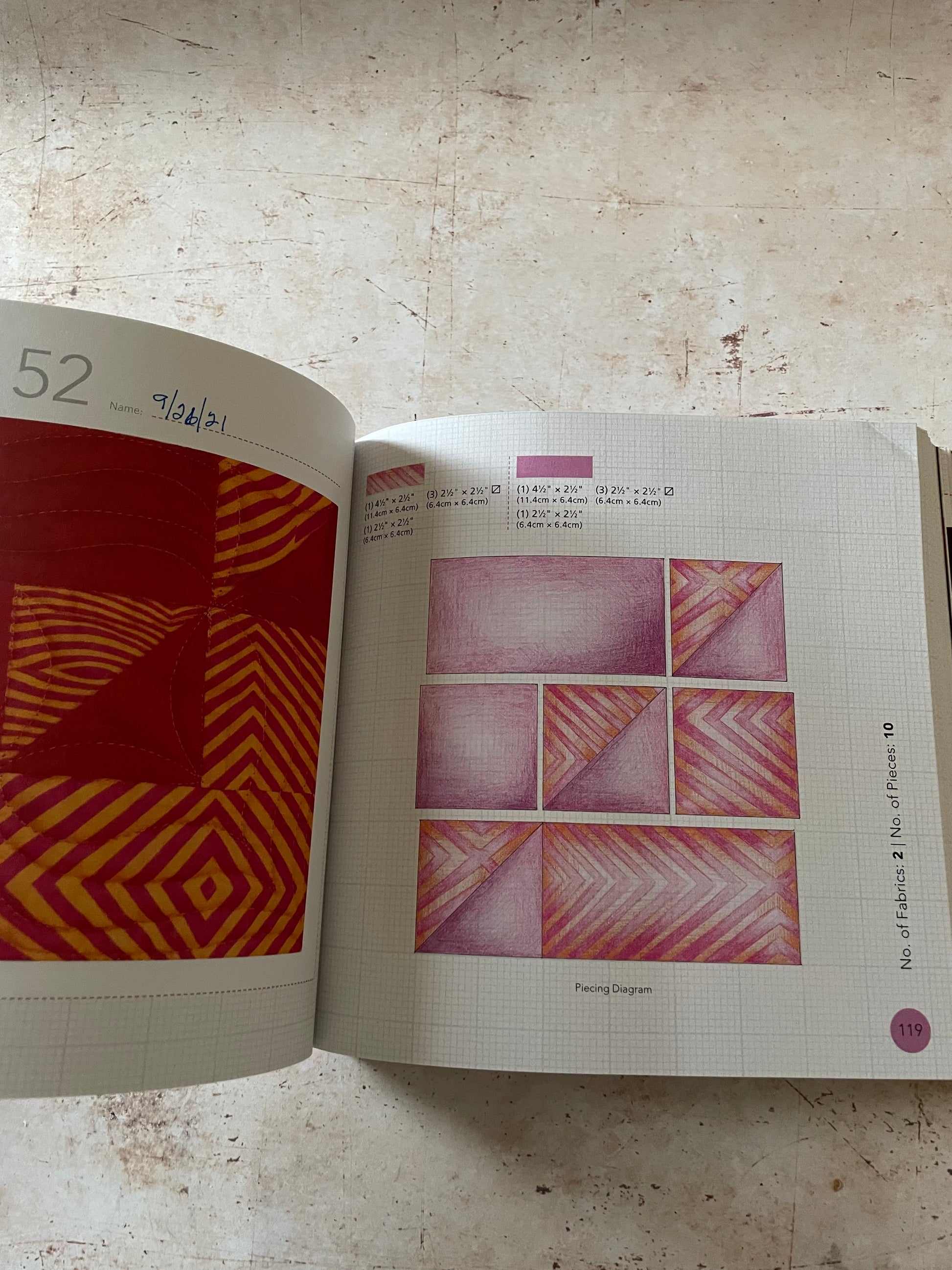 An open quilt pattern book lying on a textured beige surface, showing two pages with geometric fabric designs in red, orange, pink, and purple. 