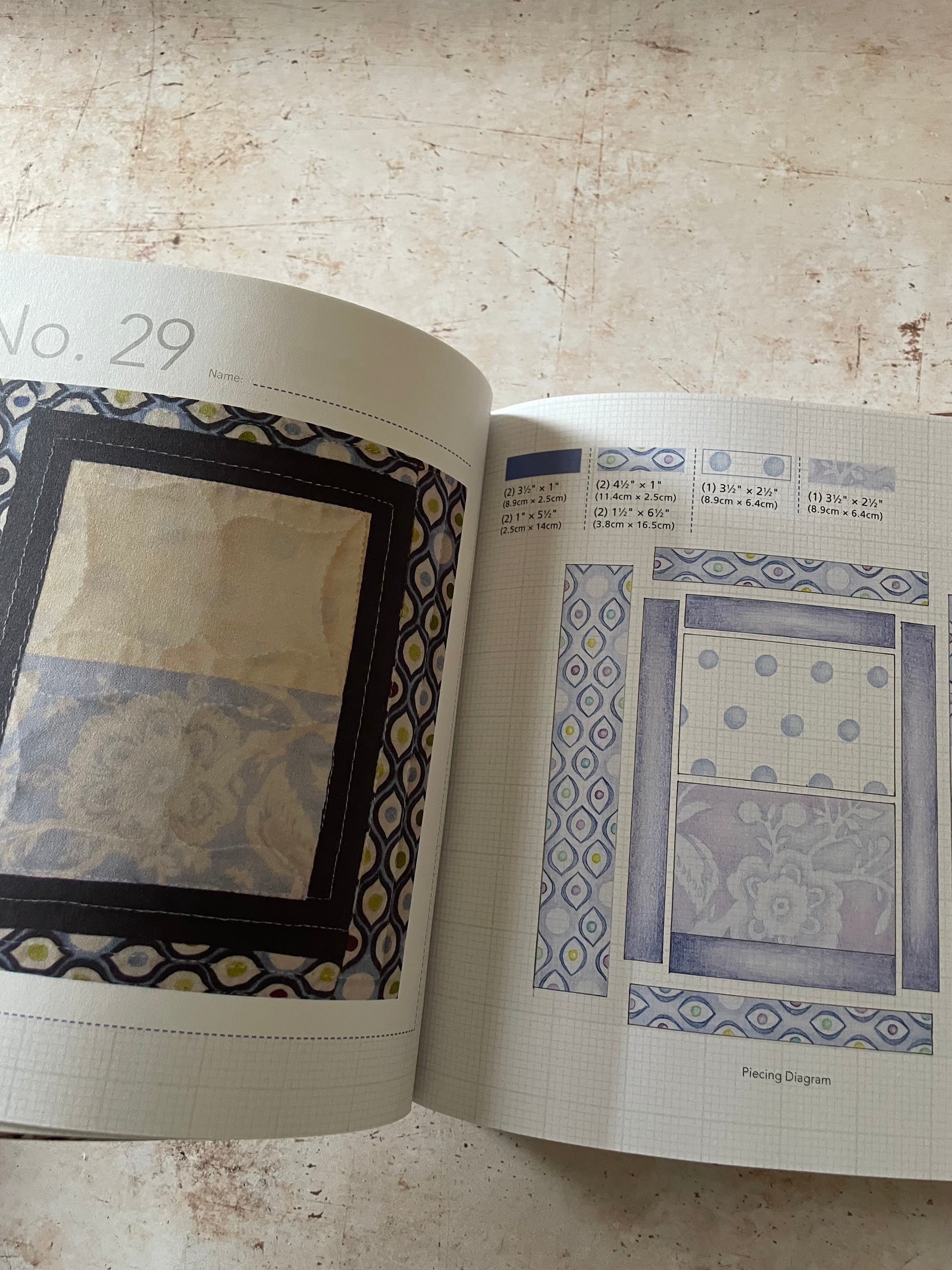 An open book showing printed fabric  pattern pages with blue, gray, and beige designs, photographed on a textured beige surface.