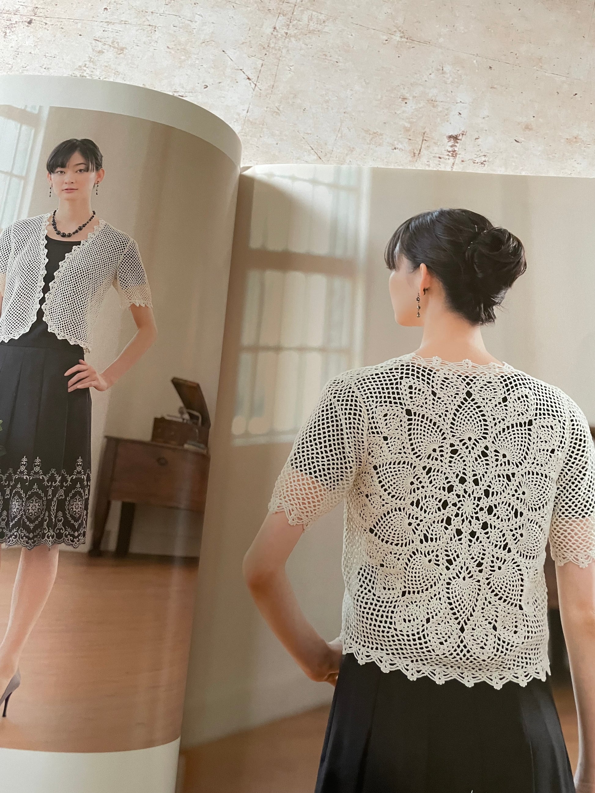 A woman shown from the back wearing a white crocheted short-sleeve cardigan with an intricate lace pattern, paired with a black skirt. 