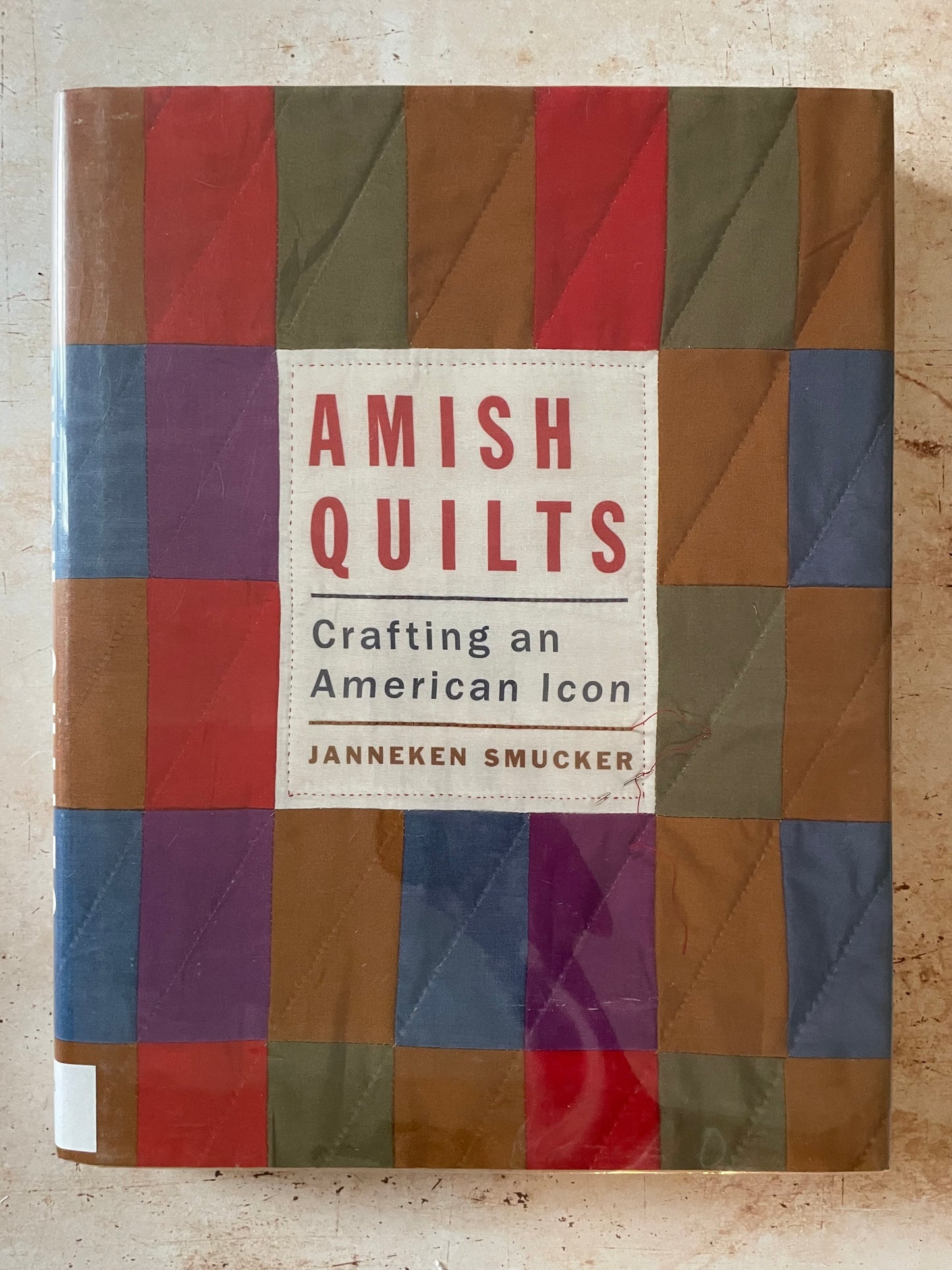 The book cover of 'Amish Quilt: Crafting an American Icon' by Janneken Smucker, featuring a colorful patchwork quilt design with a central cream-colored title block.